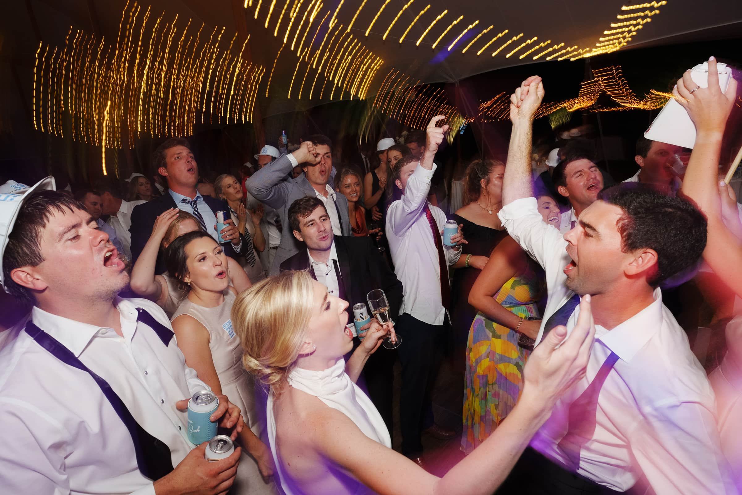 dunn wedding photography - reaves & zach A group of people having fun at a lively party, with some holding drinks and singing, as captured by Dunn wedding photography. Festive string lights create a warm, dynamic atmosphere, perfectly framing every joyful smile. Everyone seems to be enjoying the celebratory mood.