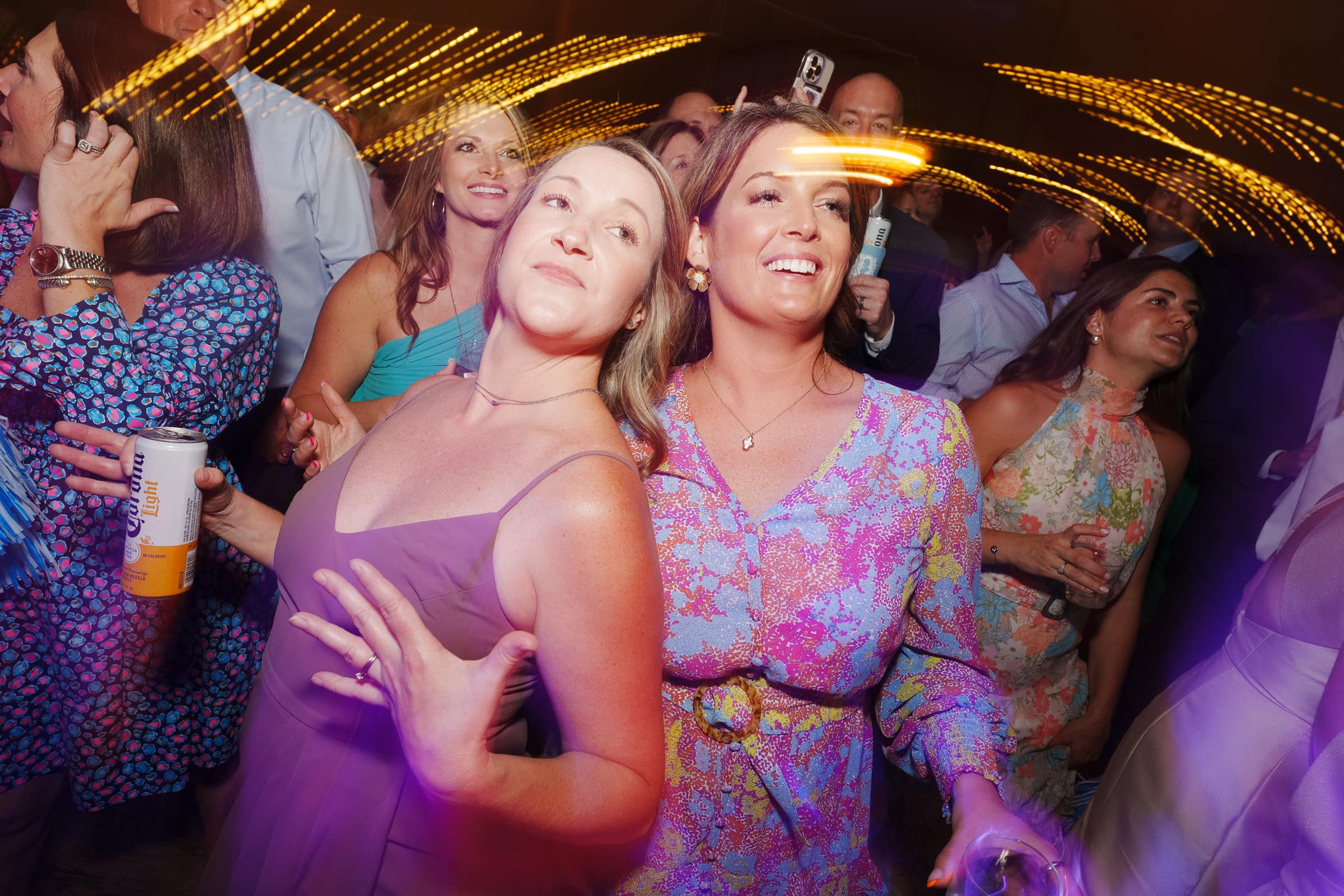 dunn wedding photography - reaves & zach A group of people enjoy dancing at a lively party, captured beautifully by Dunn wedding photography. The scene is full of energy, with a mix of colors and motion blurs from lights overhead. Two women in the foreground look happy and engaged in the moment, holding drinks and smiling.