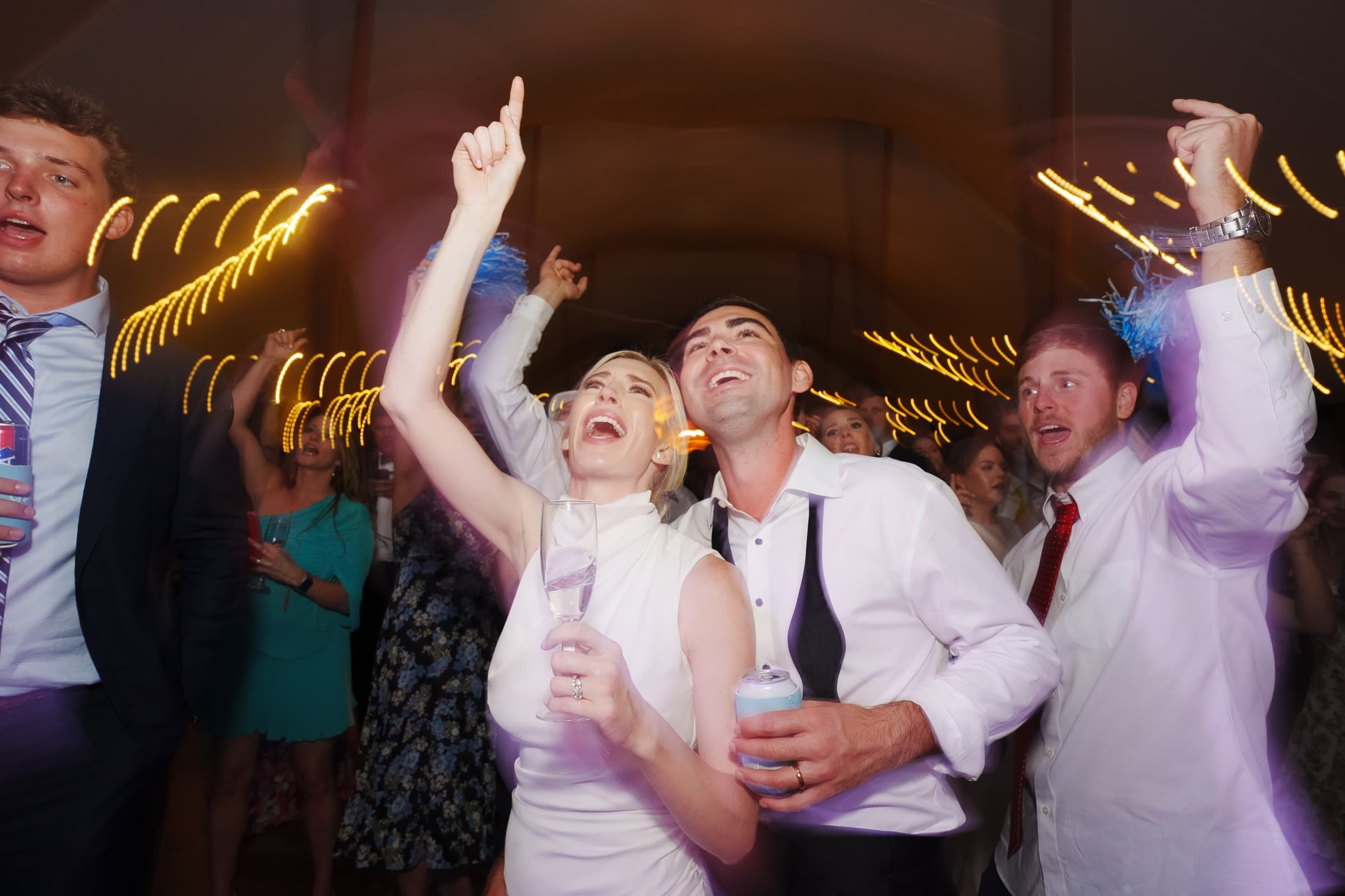 dunn wedding photography - reaves & zach At what appears to be a Dunn wedding photography session, a group of people dressed in formal attire are dancing and celebrating. They appear joyful, with some raising their hands and holding drinks. The background shows blurred, warm string lights, suggesting a festive atmosphere.
