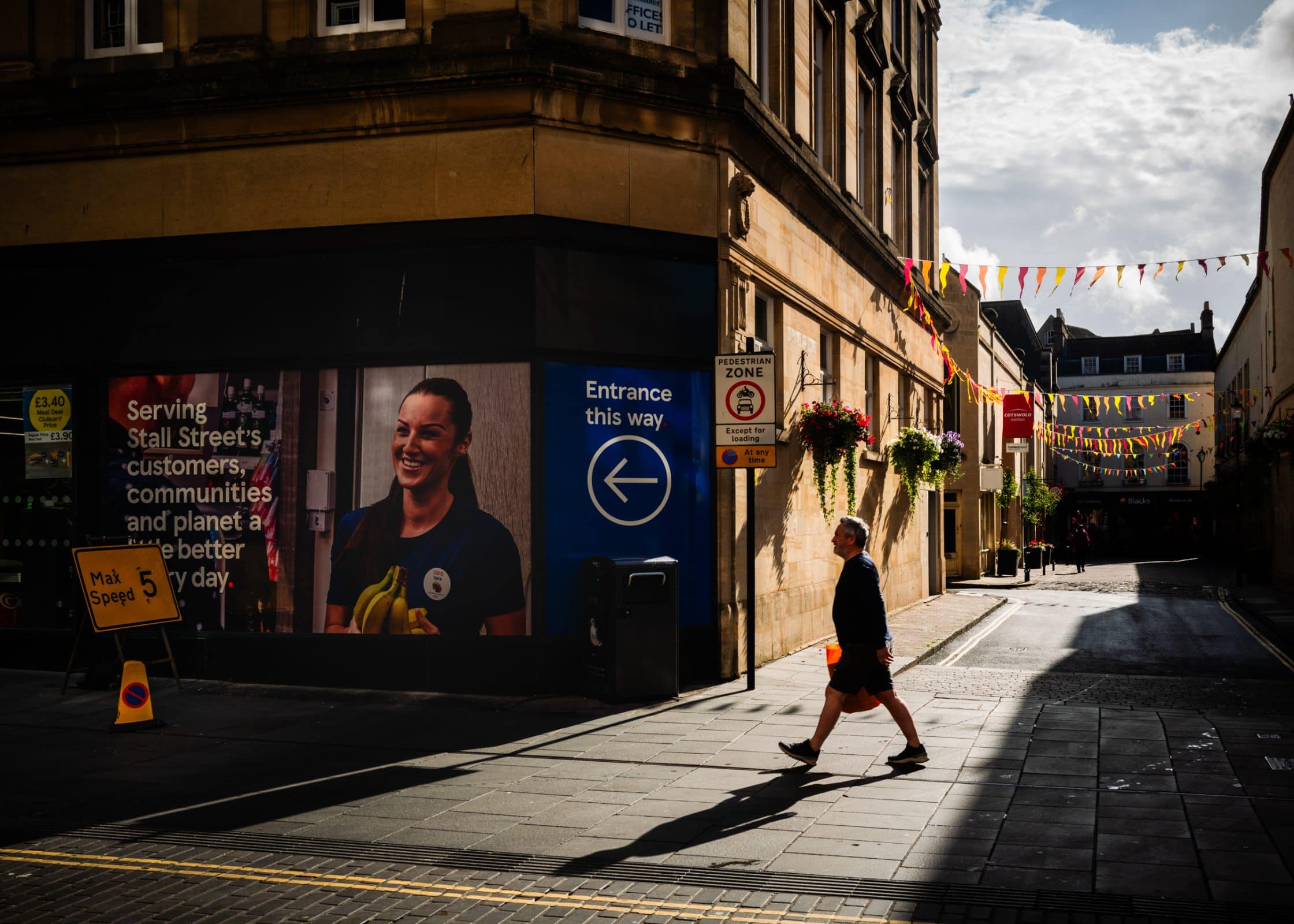 strengthening my photography skillset: a journey of self-development A person with a keen photography skillset strolls along a sunlit street past a building adorned with colorful bunting overhead. A large sign on the building reads, Serving Stall Streets customers, communities, and planet a little better every day, with an arrow pointing to the entrance on the right.