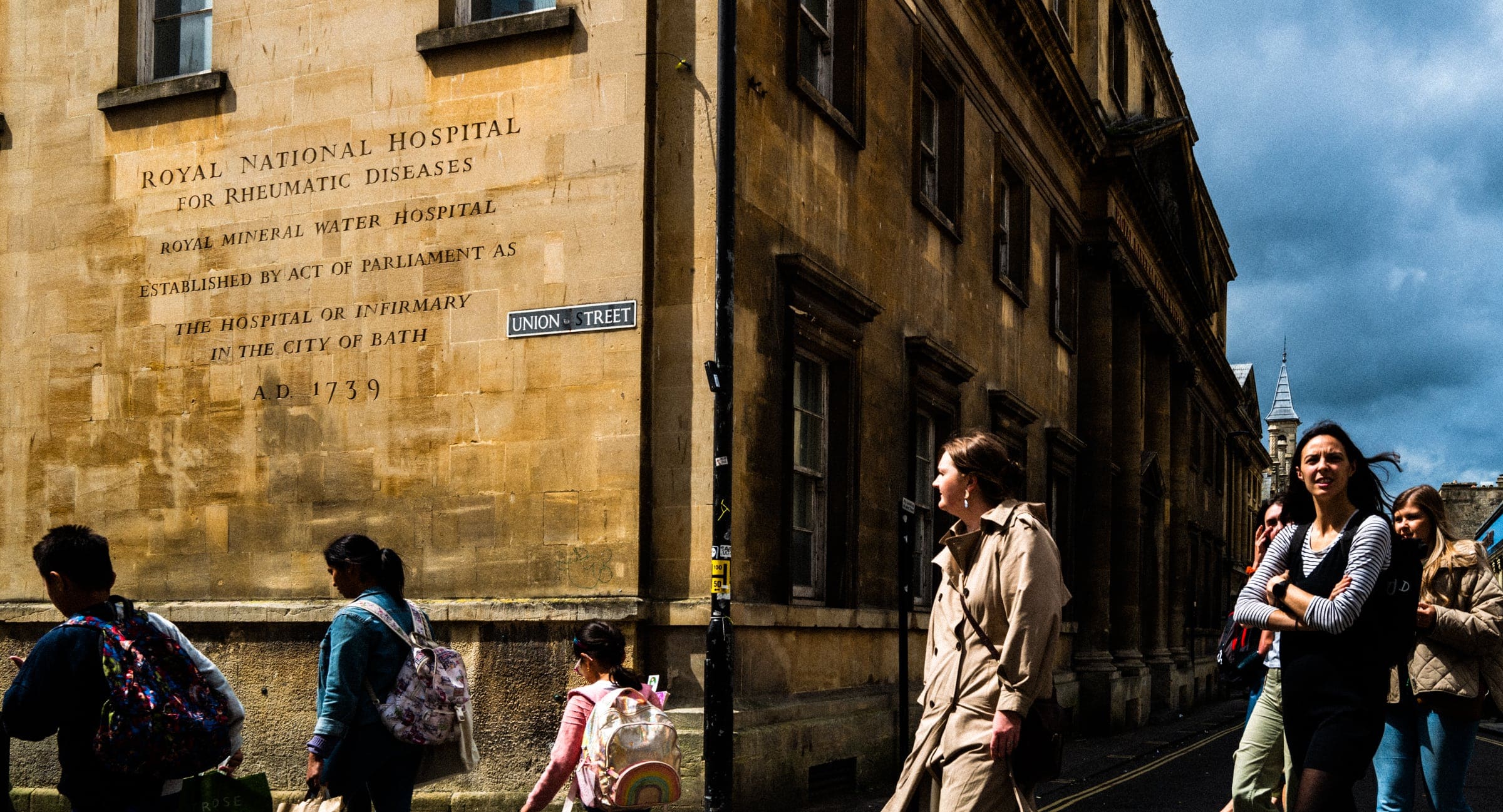 strengthening my photography skillset: a journey of self-development People with a keen photography skillset capture the bustling street as pedestrians walk past a hospital building. The wall displays engraved text about the Royal National Hospital for Rheumatic Diseases. With partly cloudy skies above, the scene vividly portrays urban life.