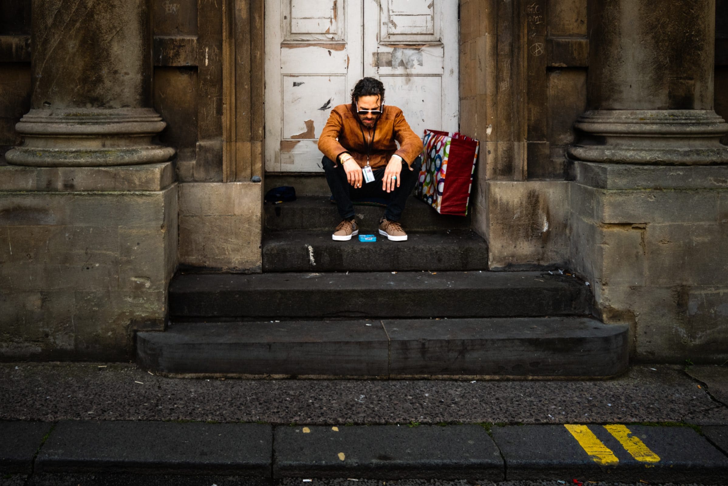 strengthening my photography skillset: a journey of self-development A person with sunglasses and a brown jacket sits gracefully on steps outside a weathered building, showcasing their photography skillset with an artfully placed colorful bag and red luggage. The scene captures urban charm, framed by a closed wooden door and bold yellow street lines.