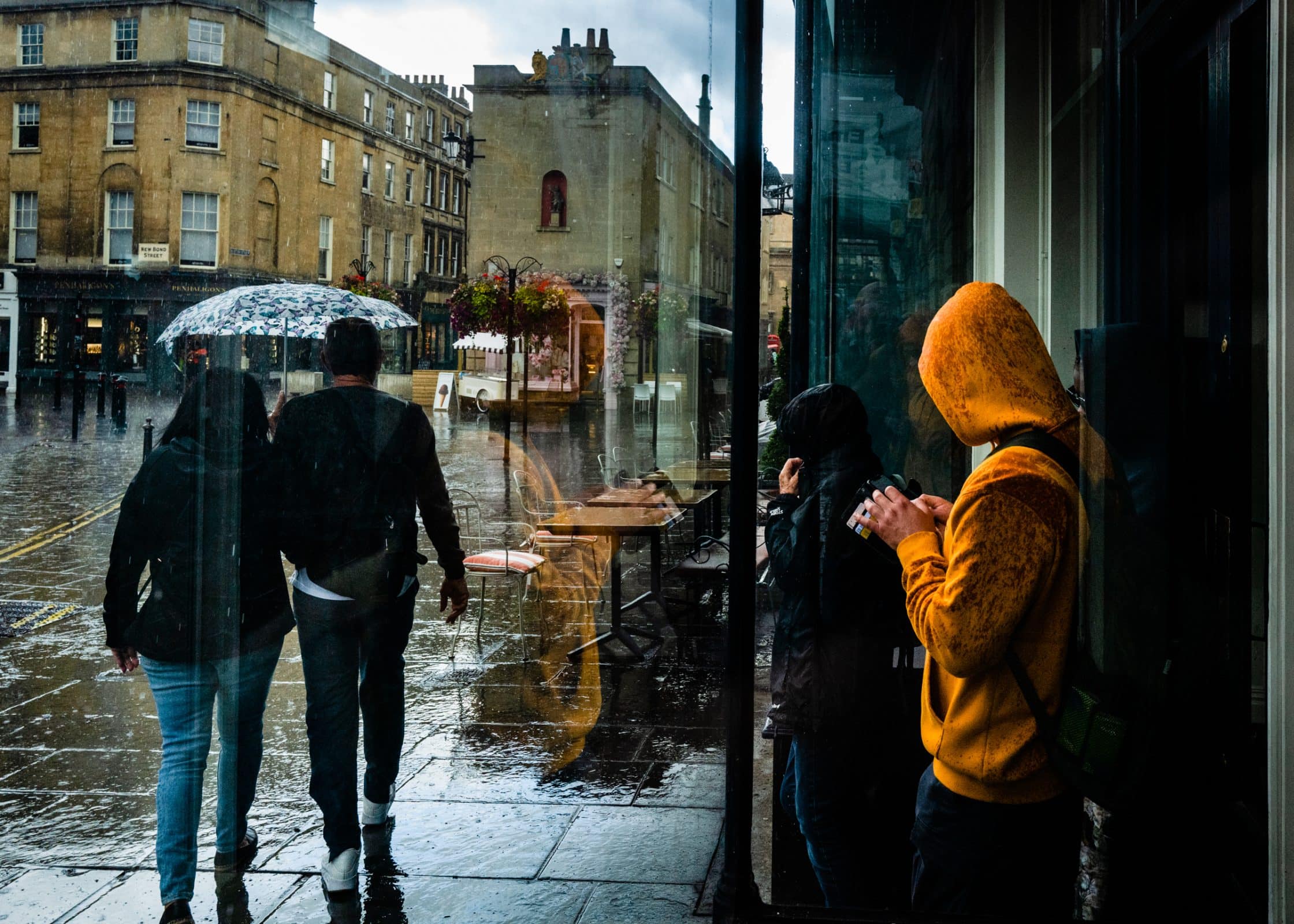 strengthening my photography skillset: a journey of self-development People walking on a rainy street, some with umbrellas. A person in an orange hoodie stands under a shelter, using a smartphone. With expert photography skillset, one captures the wet pavement reflecting the buildings and street scene in stunning detail.