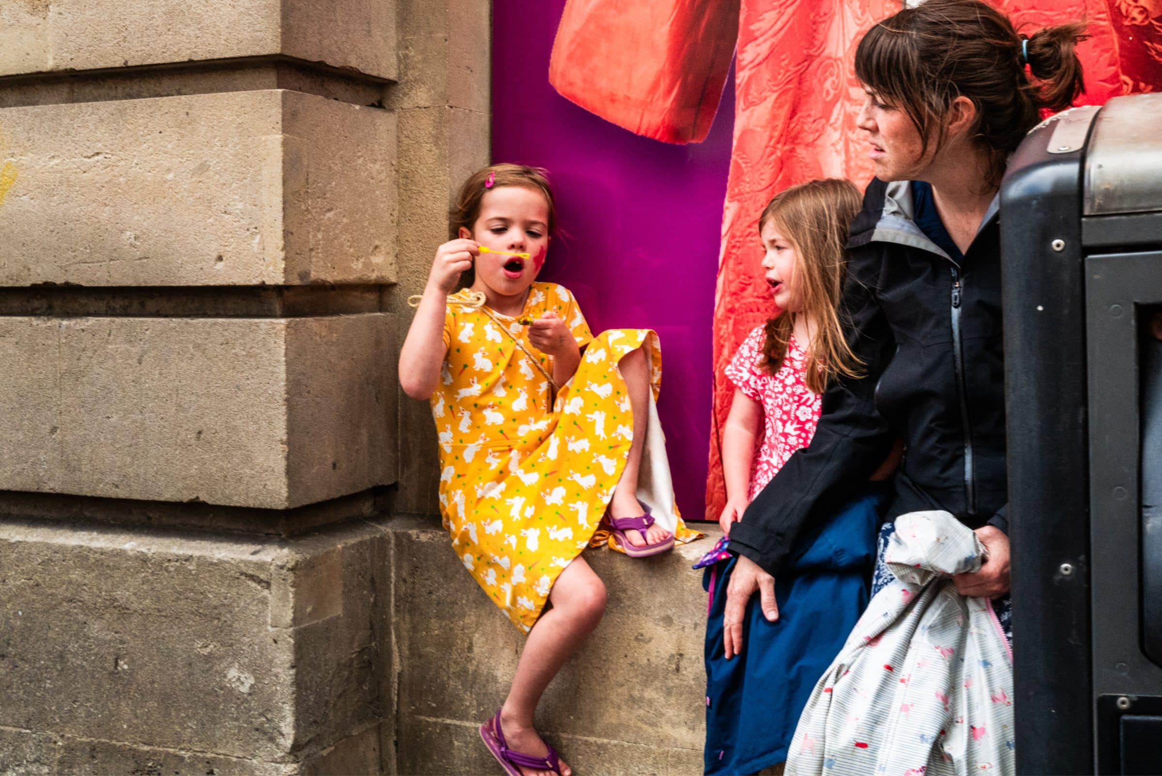 strengthening my photography skillset: a journey of self-development A child in a yellow dress expertly blows bubbles, showcasing a photography skillset, while sitting on a ledge next to a girl in red and a woman with a jacket. They are framed against a stone wall with colorful fabric behind them on this bright day.