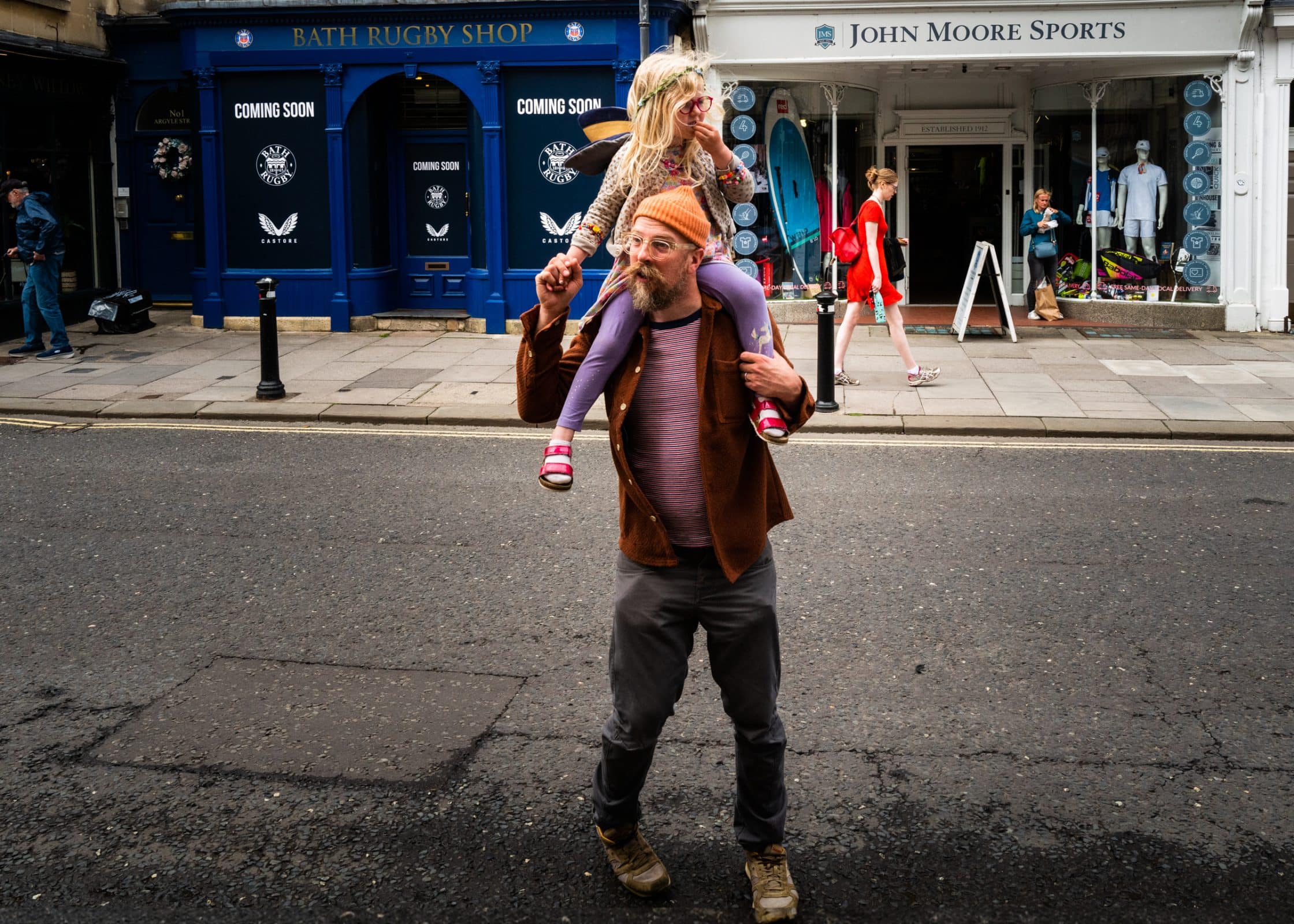 strengthening my photography skillset: a journey of self-development A man showcasing his photography skillset captures a moment while wearing a brown jacket and orange hat, as he carries a child in a pink outfit with a snack stick on his shoulders across the street. Shops can be seen bustling in the background.