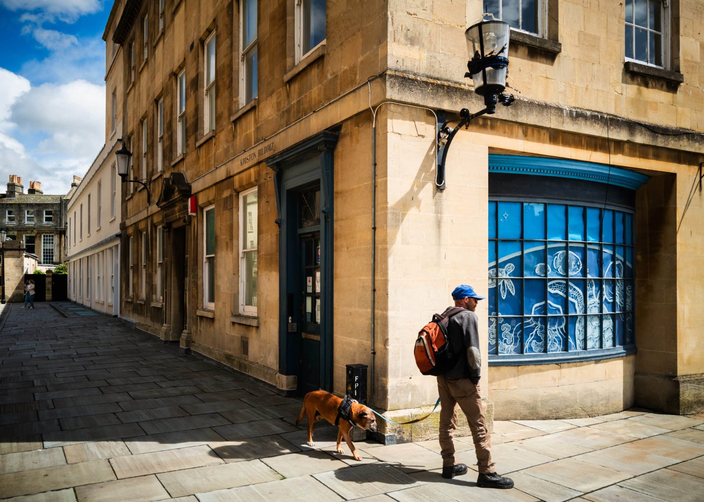 strengthening my photography skillset: a journey of self-development A person showcasing their photography skillset captures a moment of walking a brown dog on a leash beside a historic stone building. The scene features a curved blue-painted window displaying decorative art, with the stone-paved street beneath a partly cloudy sky.