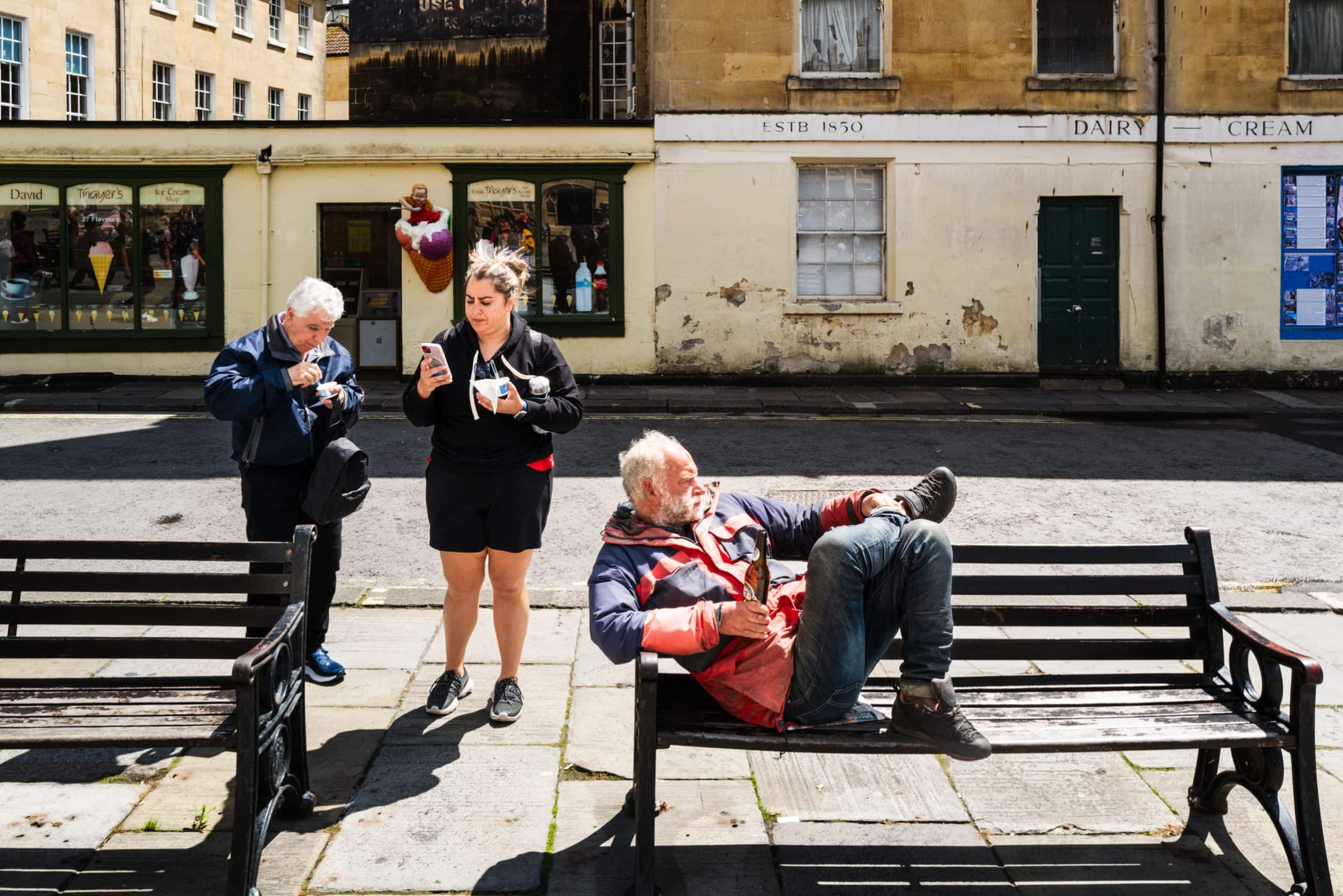 strengthening my photography skillset: a journey of self-development Three people in a sunny street scene showcase their photography skillset. One man is reclining on a bench, while a woman stands nearby holding a phone and an ice cream. Another man stands beside her, focused on something in his hands. Historic buildings are in the background.