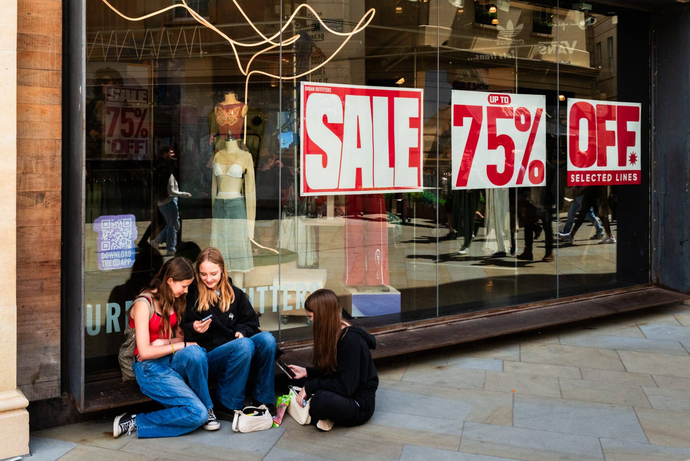 strengthening my photography skillset: a journey of self-development Three young people sit on the ground outside a store with bold red signs reading SALE and 75% OFF Selected Lines. Engrossed in their phones, they ignore the mannequins nearby—a scene capturing urban life that truly tests any photographer's skillset.