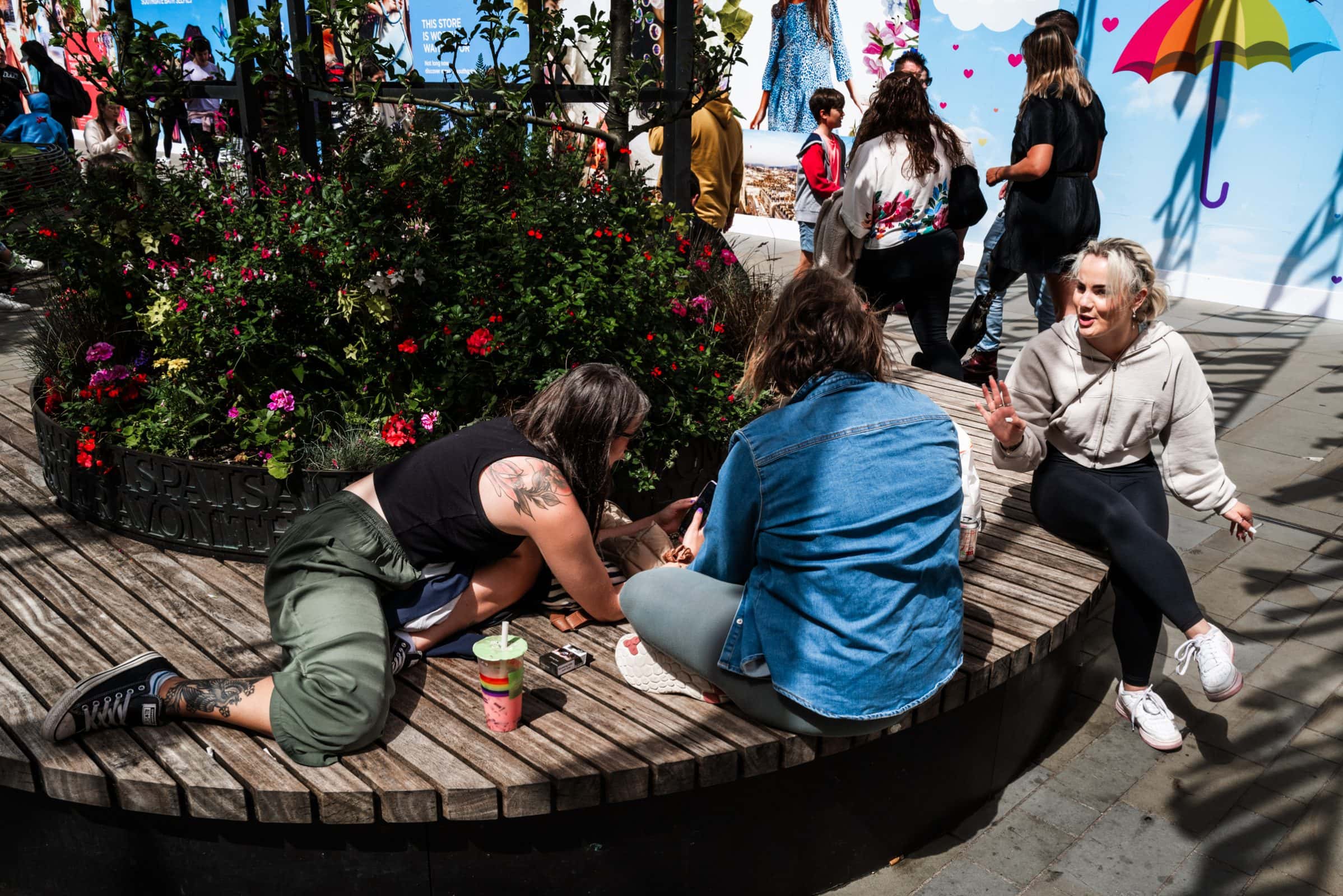 strengthening my photography skillset: a journey of self-development Three friends, showcasing their photography skillset, sit on a round wooden bench surrounded by flowers, chatting and laughing on a sunny day. People walk by in the background against a colorful mural featuring an umbrella and clouds.
