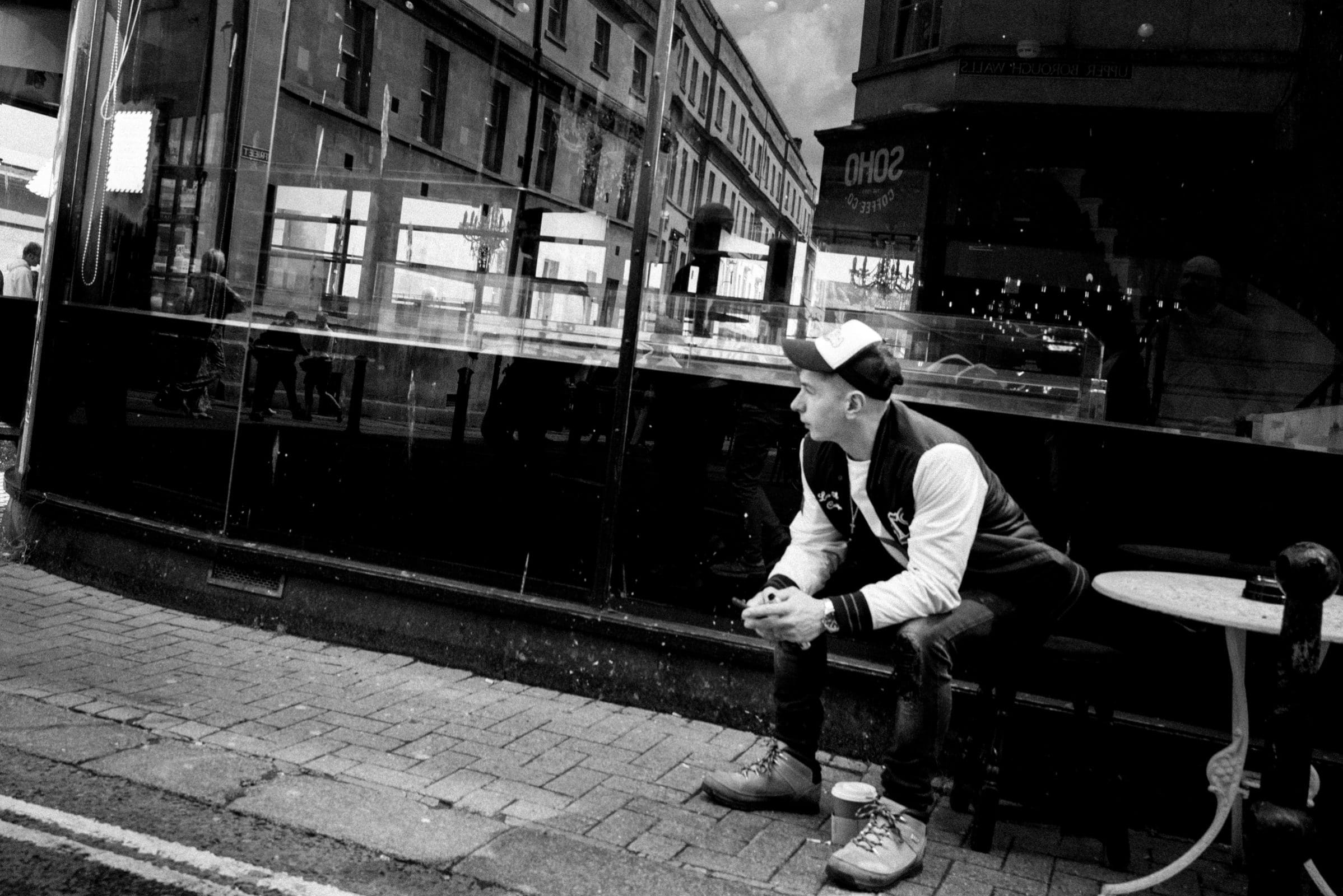 strengthening my photography skillset: a journey of self-development Black and white photo showcasing photography skillset: a man sits outside a café on a city street, wearing a baseball cap and jacket, gazing to his right. The reflection of buildings and pedestrians is artfully captured in the large window behind him.