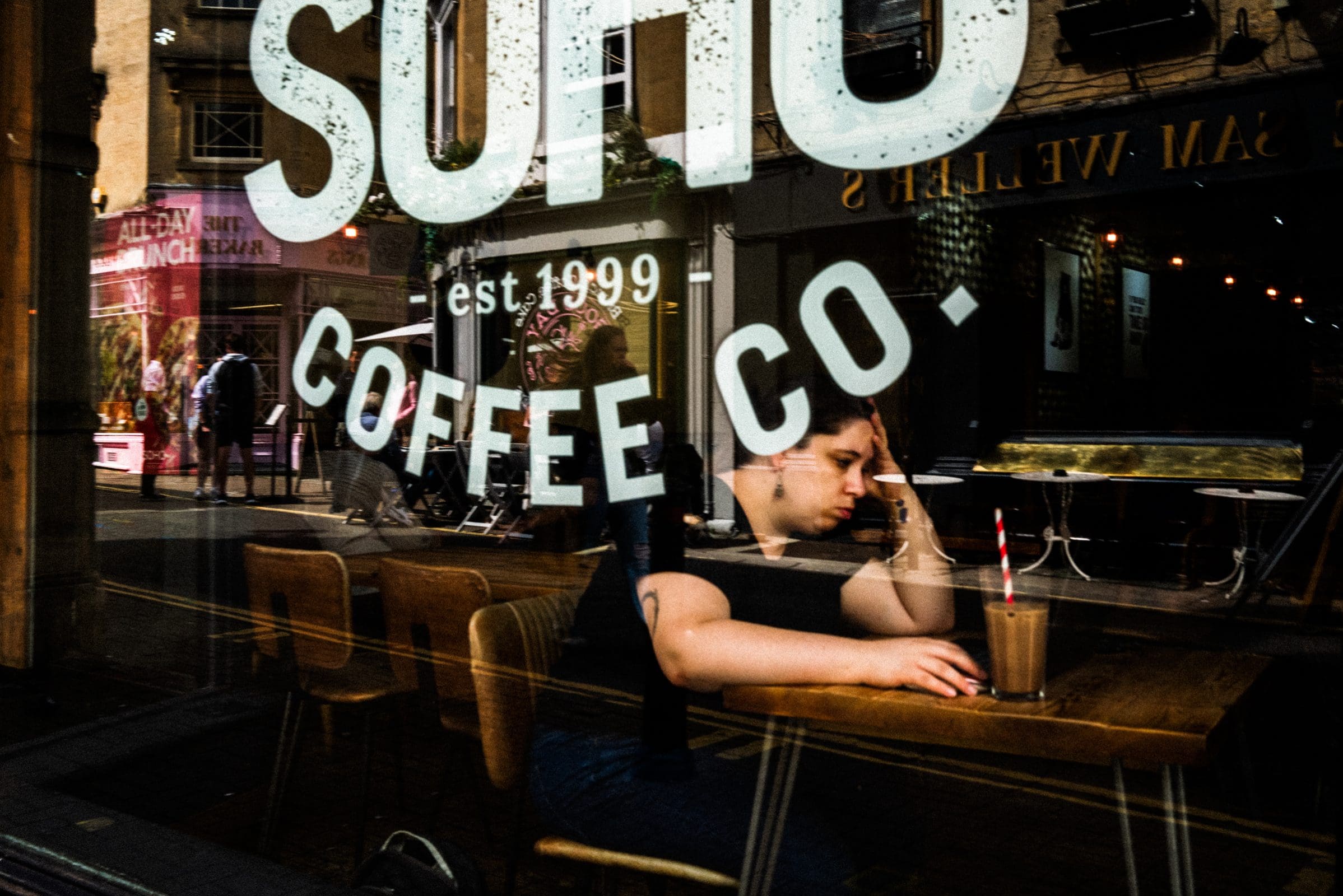 strengthening my photography skillset: a journey of self-development A person, showcasing keen photography skillset, sits inside a cafe, leaning on the table with a drink in hand. The cafe's large window reflects a busy street outside, where bold SOHO COFFEE CO. lettering stands out on the glass.
