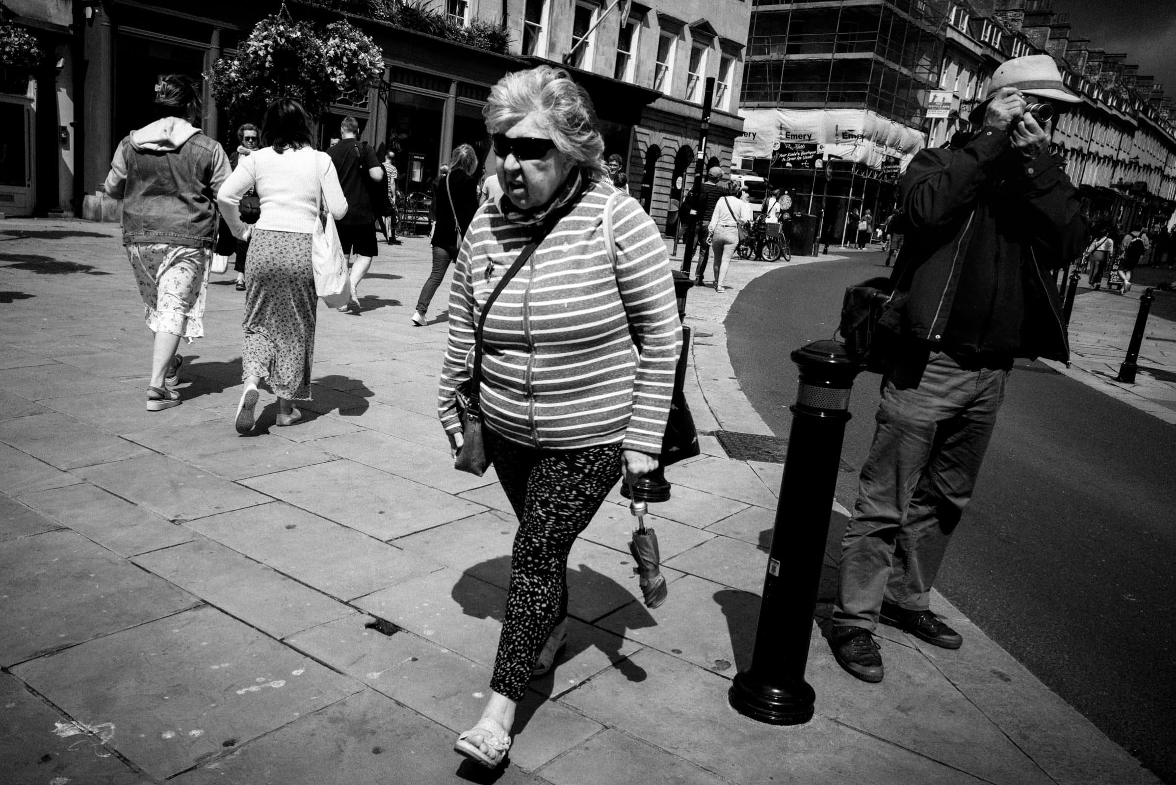 strengthening my photography skillset: a journey of self-development A black-and-white street scene captures a woman walking on a sidewalk with an umbrella. Nearby, a man showcases his photography skillset by looking through a camera. Other pedestrians and buildings line the street under the partly cloudy sky.