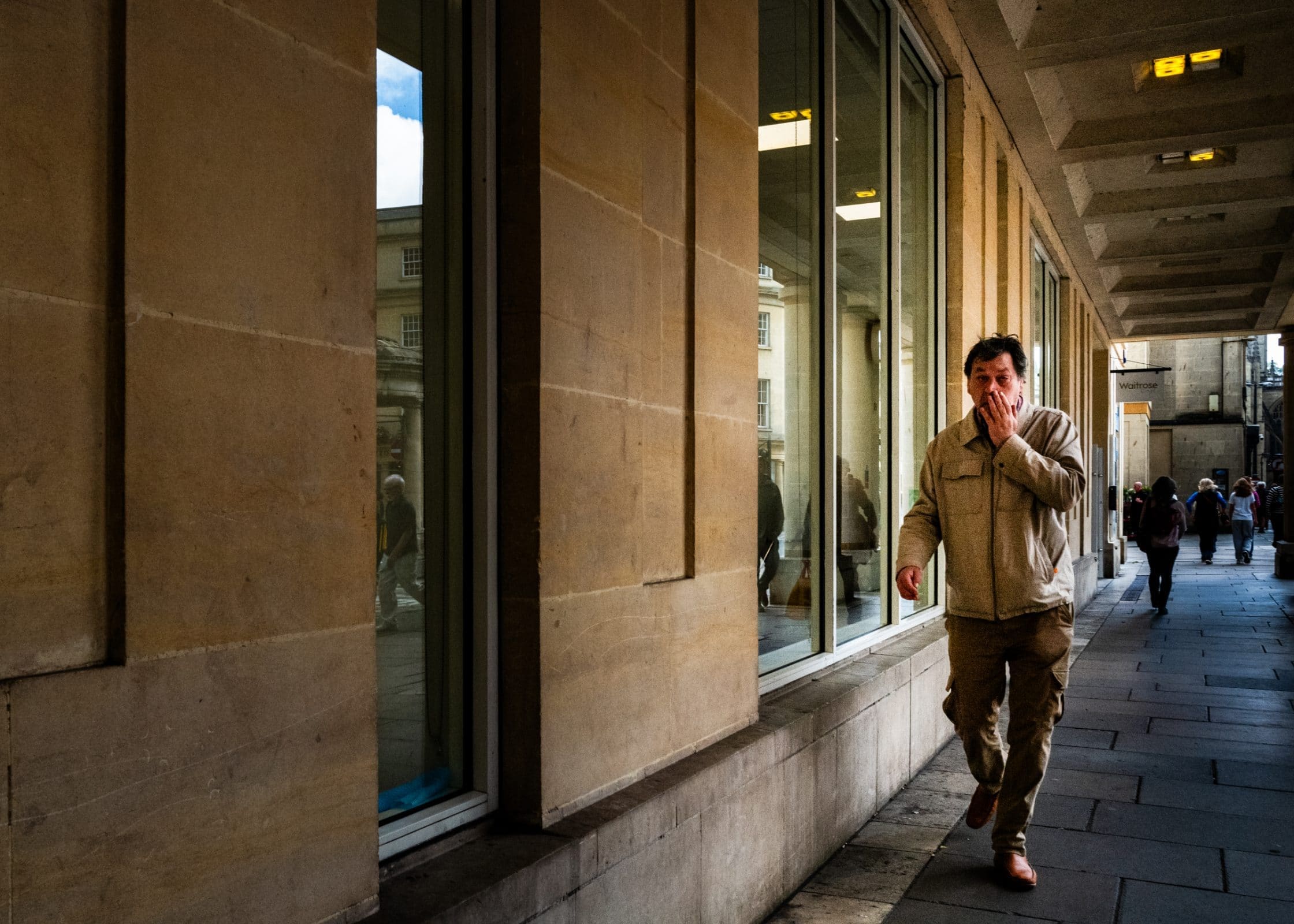 strengthening my photography skillset: a journey of self-development A man in a beige jacket strolls along a sidewalk by a building with large windows, capturing the scene with his eye for photography skillset. He's touching his face, and others walk in the distance. Overhead lights illuminate the partially cloudy sky above.