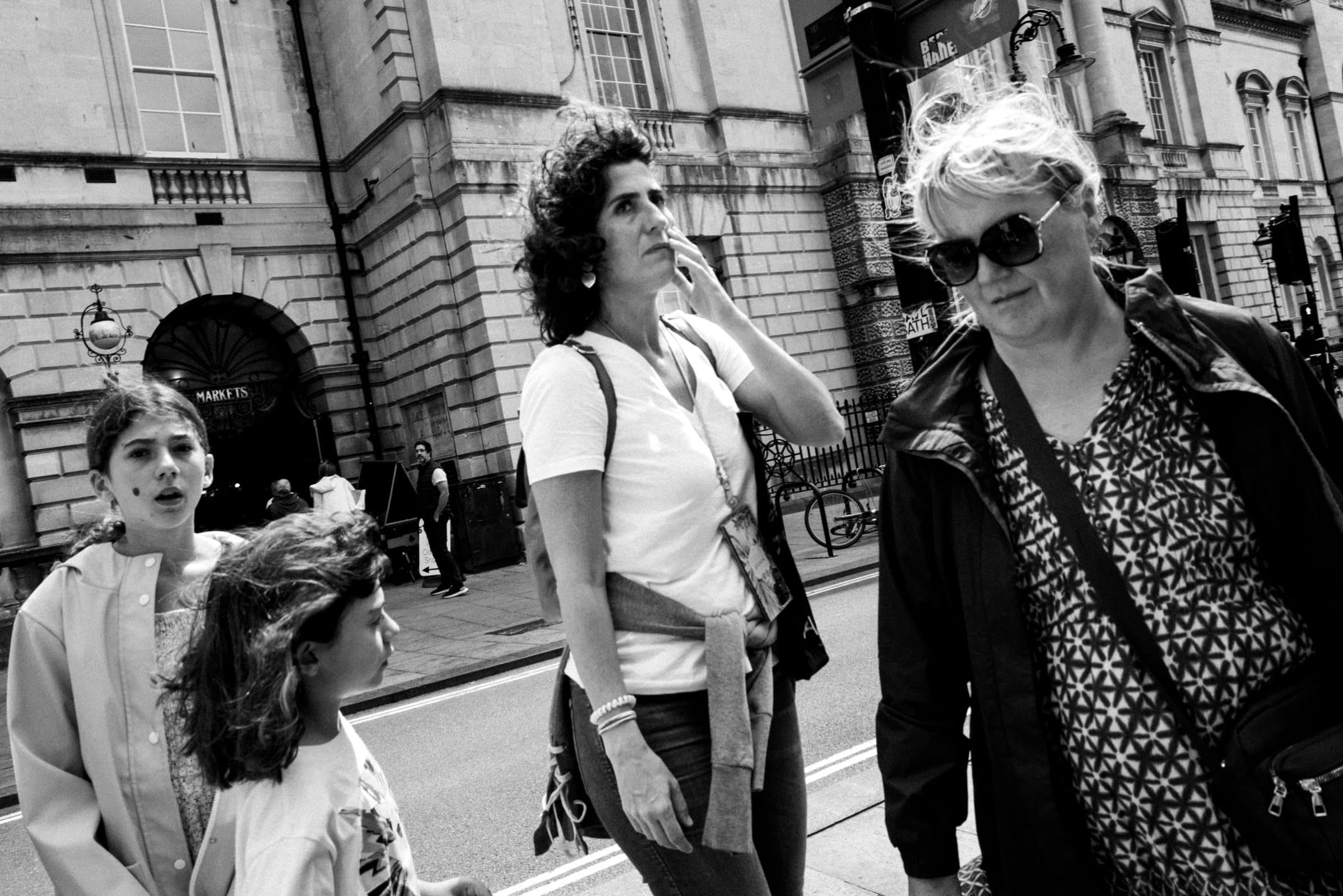 strengthening my photography skillset: a journey of self-development A black and white photo showcasing impressive photography skillset captures four people walking on a city street. Two women, one with curly hair and the other in sunglasses, stroll together, while two young girls are nearby. The backdrop reveals a historic building.