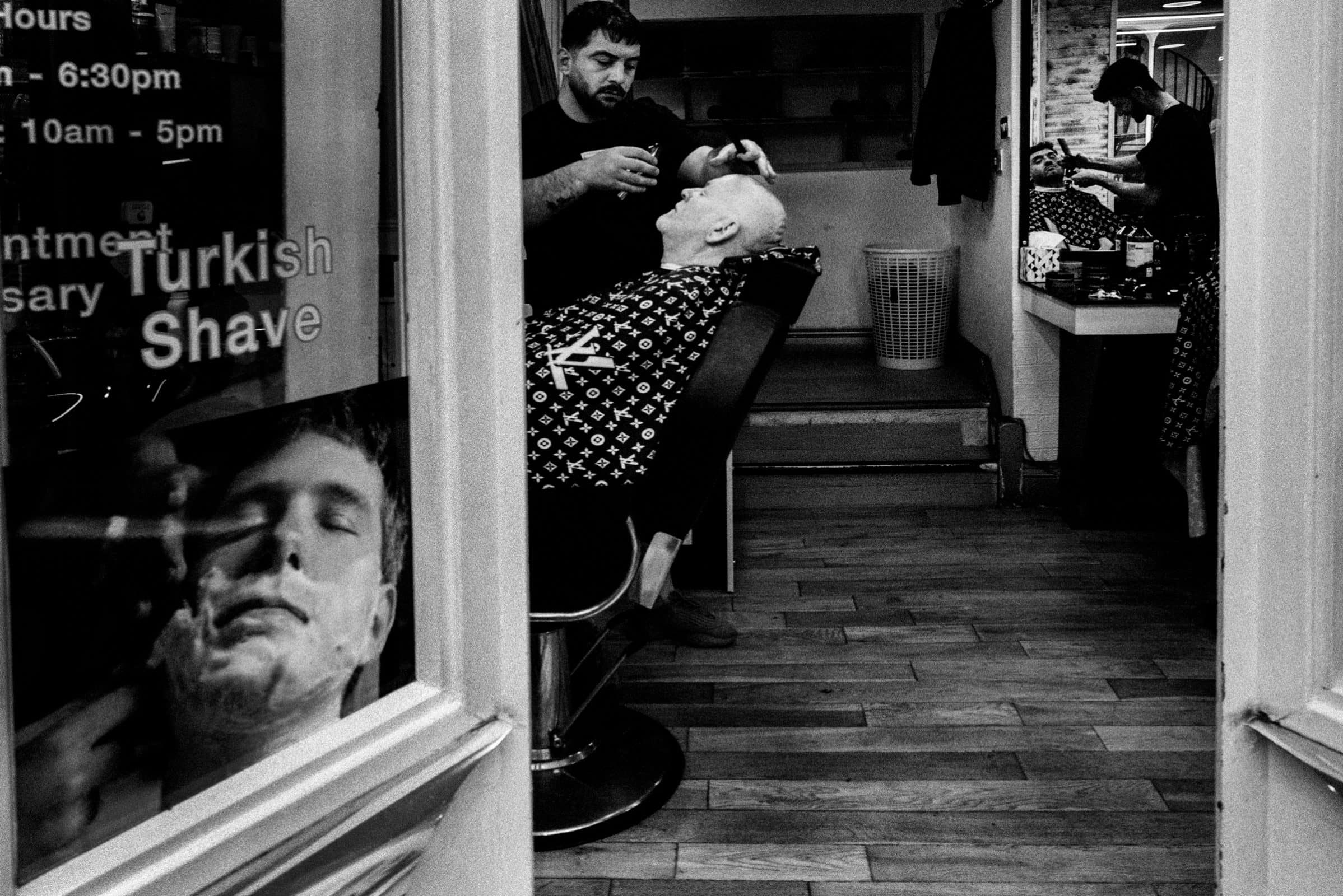 A barber expertly shaves a seated customer with a straight razor, showcasing his photography skillset in capturing the timeless scene. Wooden floors add warmth to the shop, while a mirror reflects another customer being attended to. A poster on the glass door advertises a Turkish Shave.