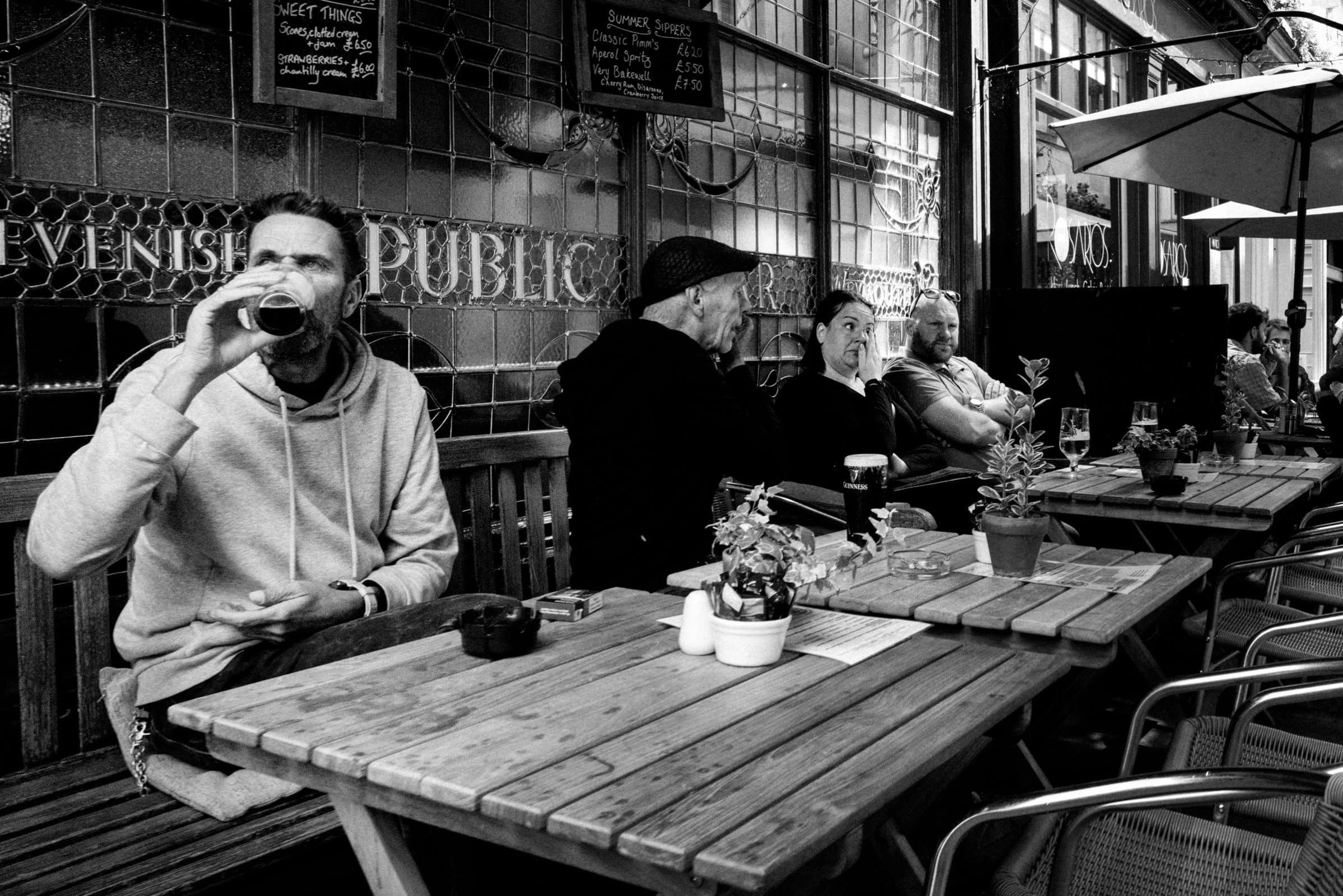 strengthening my photography skillset: a journey of self-development A black and white photo showcases several people sitting at wooden tables outside a pub. A man in the foreground drinks from a glass, while others converse in the background, demonstrating an impressive photography skillset. Plants and menu boards enhance the inviting space.