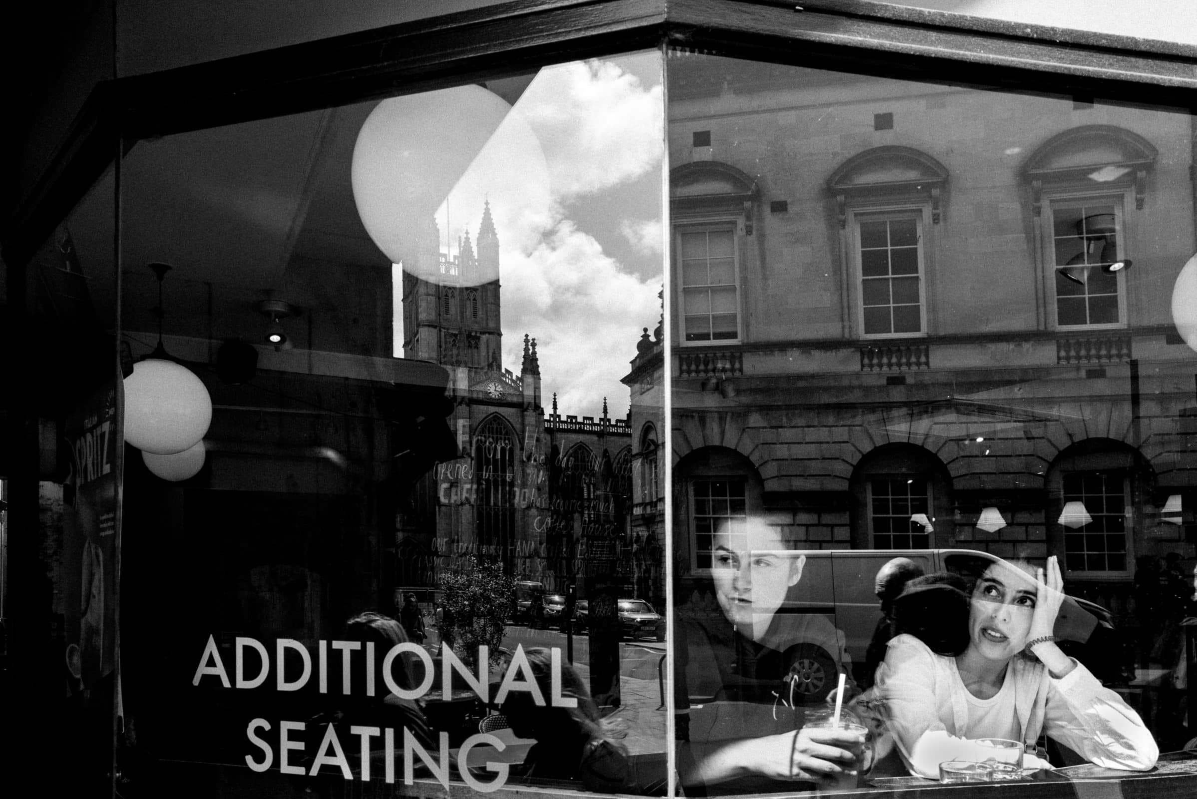 strengthening my photography skillset: a journey of self-development A black and white photo, showcasing adept photography skillset, captures two people seated inside a cafe by a window labeled "Additional Seating." Reflections of historic buildings dance outside. One person rests their head on their hand while the other holds a drink.