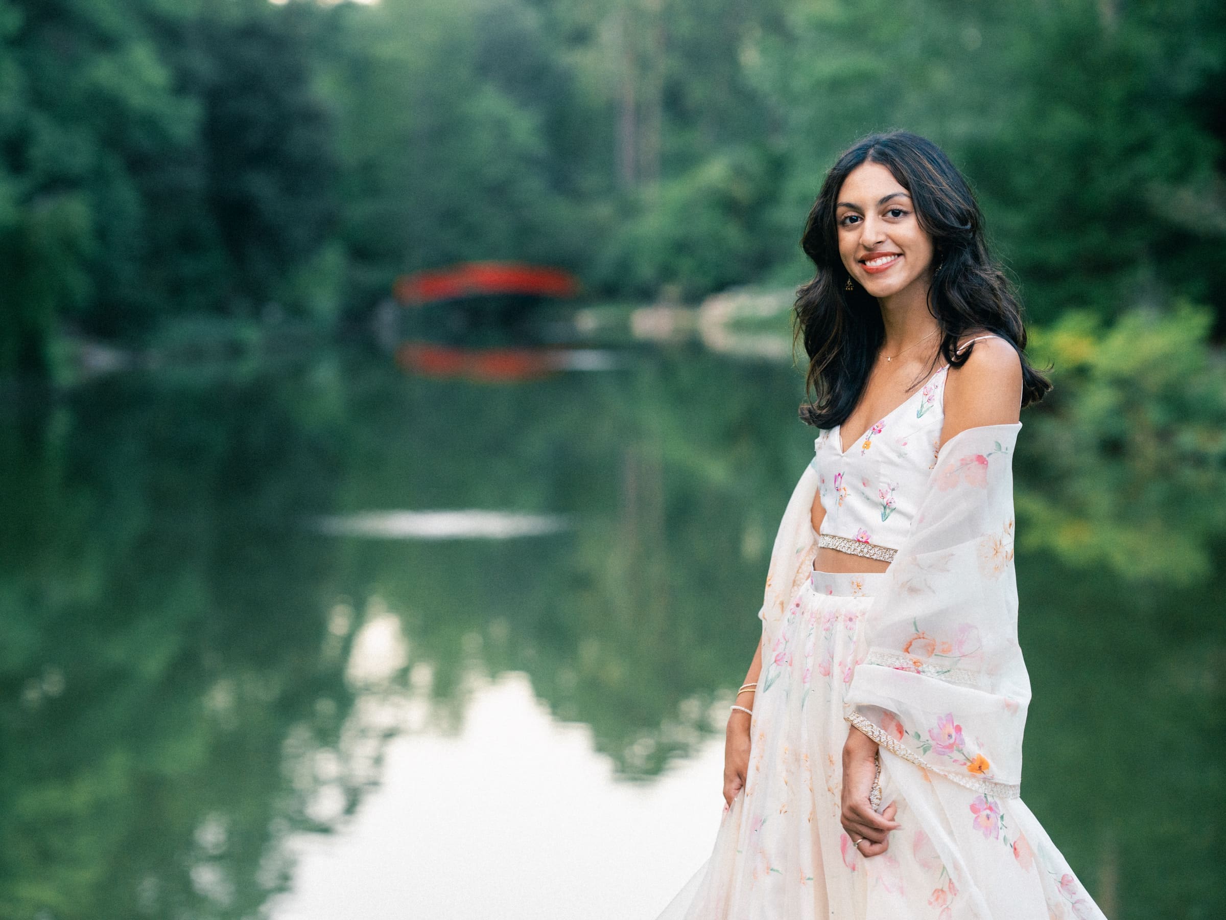 A woman in a floral dress poses for senior portraits by a serene lake, with lush greenery behind her. A red bridge is visible in the distance, mirroring beautifully in the calm water. She smiles softly, capturing the peaceful atmosphere of this picturesque setting.