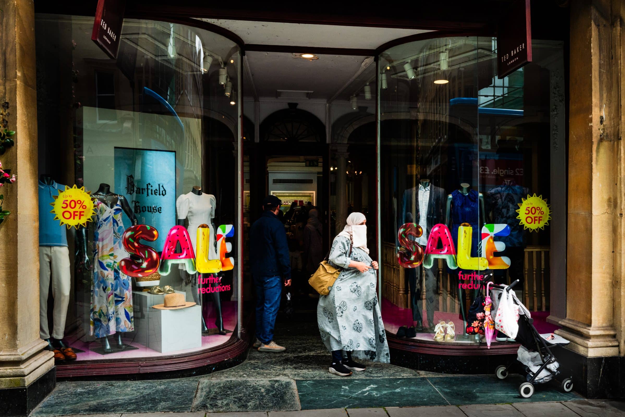 strengthening my photography skillset: a journey of self-development A person in a long grey dress and headscarf, captured with exquisite photography skillset, stands outside a clothing store adorned with SALE signs and mannequins. The shop boasts a 50% off promotion, while a stroller is parked nearby.