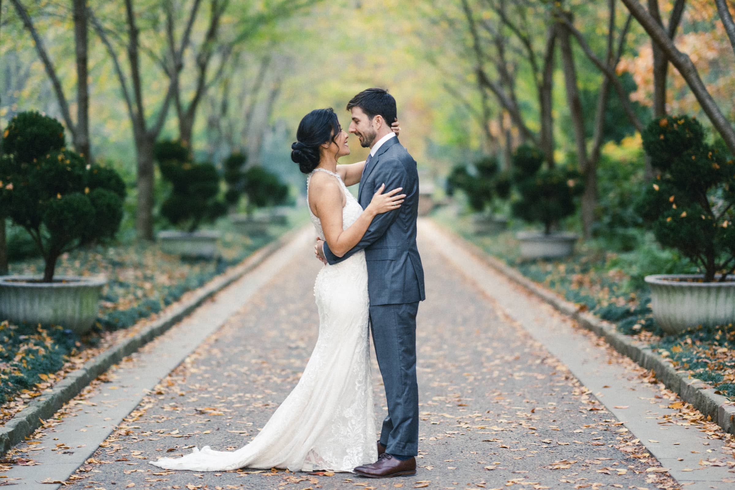 A couple in wedding attire embraces on a tree-lined path, capturing the charm of a market hall wedding. The bride wears a long, white lace gown, and the groom is in a gray suit. The surrounding trees create a serene, leafy backdrop, and the ground is strewn with fallen leaves.