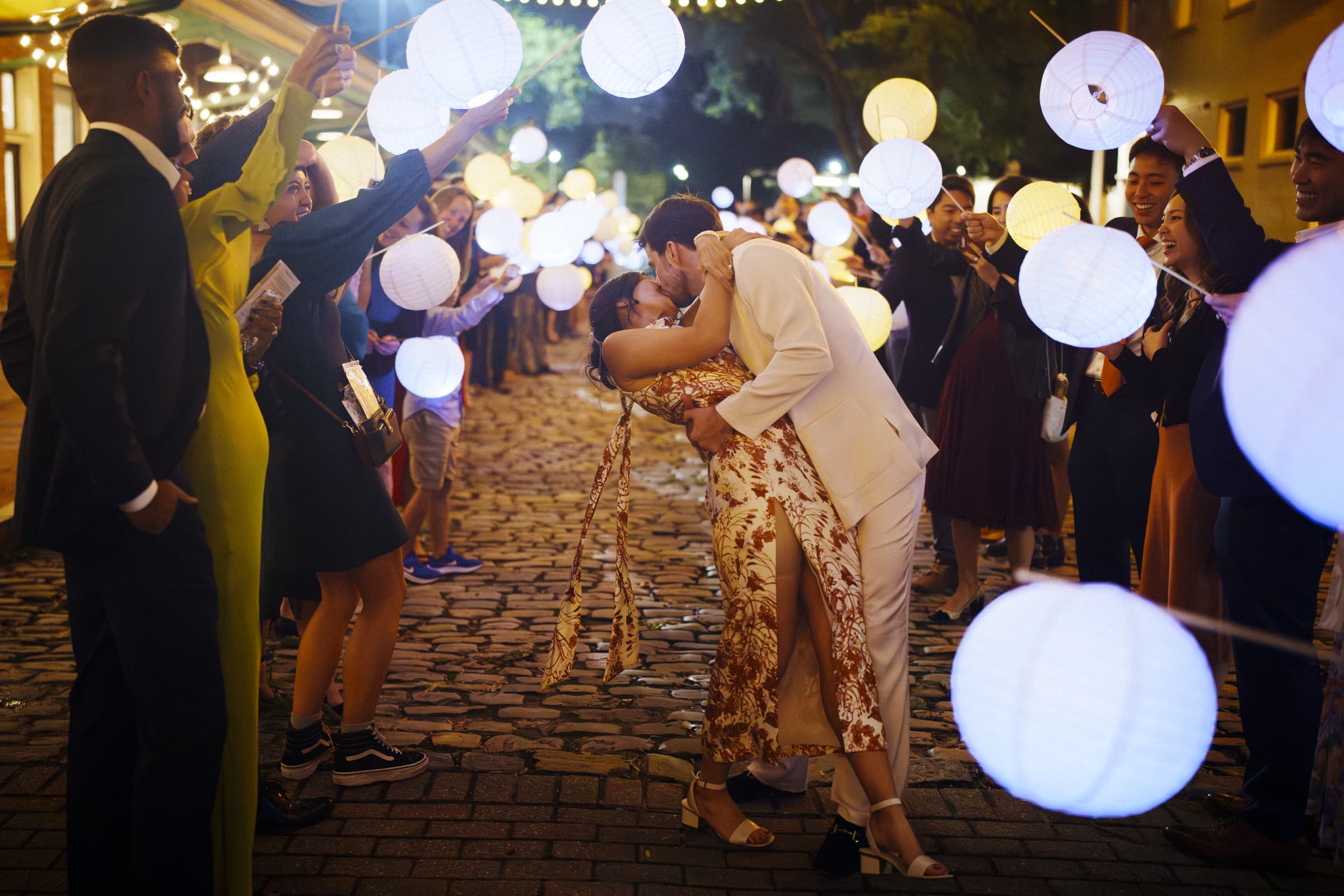 A couple in elegant attire shares a kiss under glowing lanterns held by people on a cobblestone street at night, as if stepping out from a market hall wedding, creating a festive and romantic atmosphere.