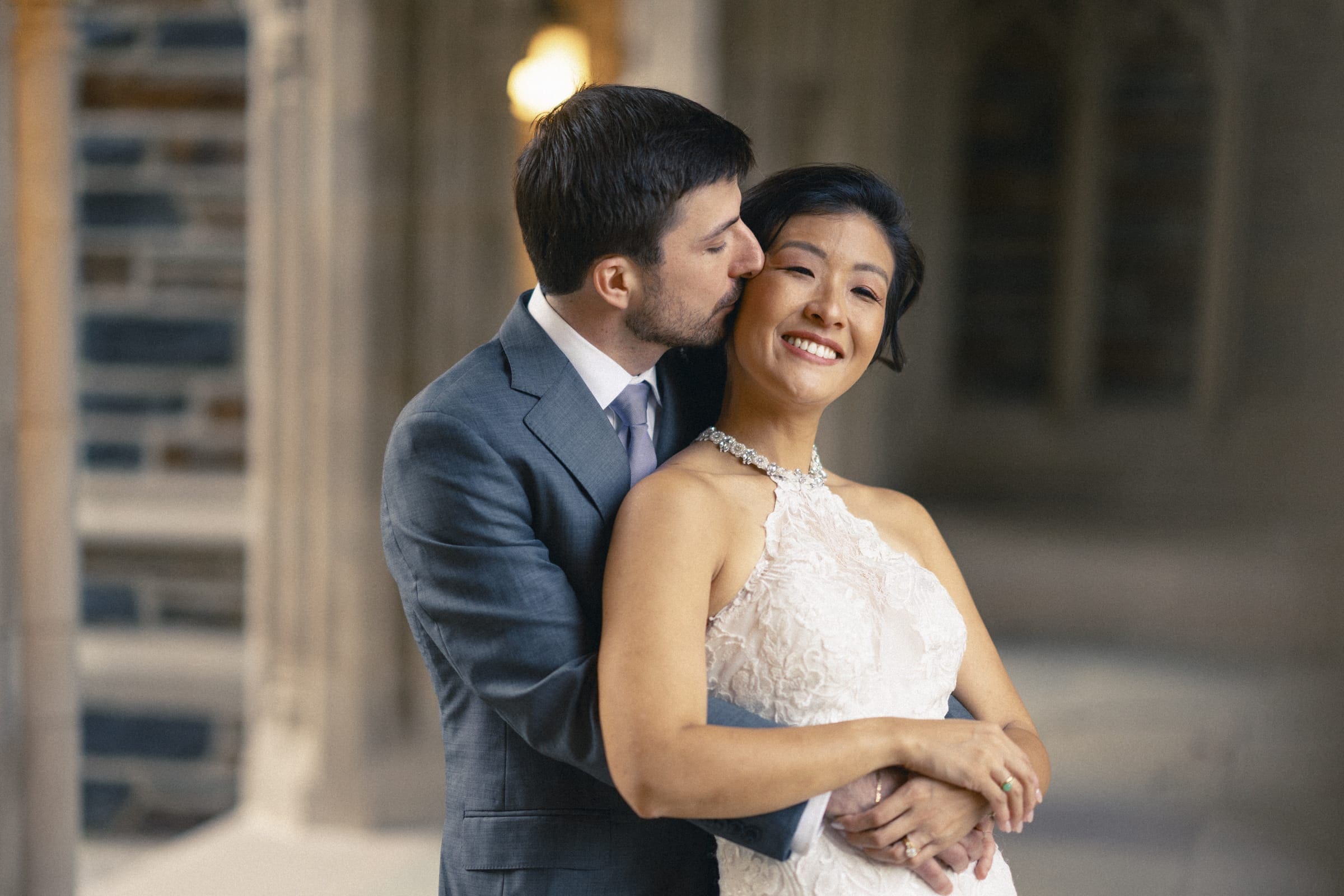 In a formal market hall setting, a couple embraces tenderly. The man, clad in a gray suit, gently kisses the woman's cheek as she smiles broadly in her white lace dress. Elegant architectural details provide a stunning backdrop to this intimate wedding moment.