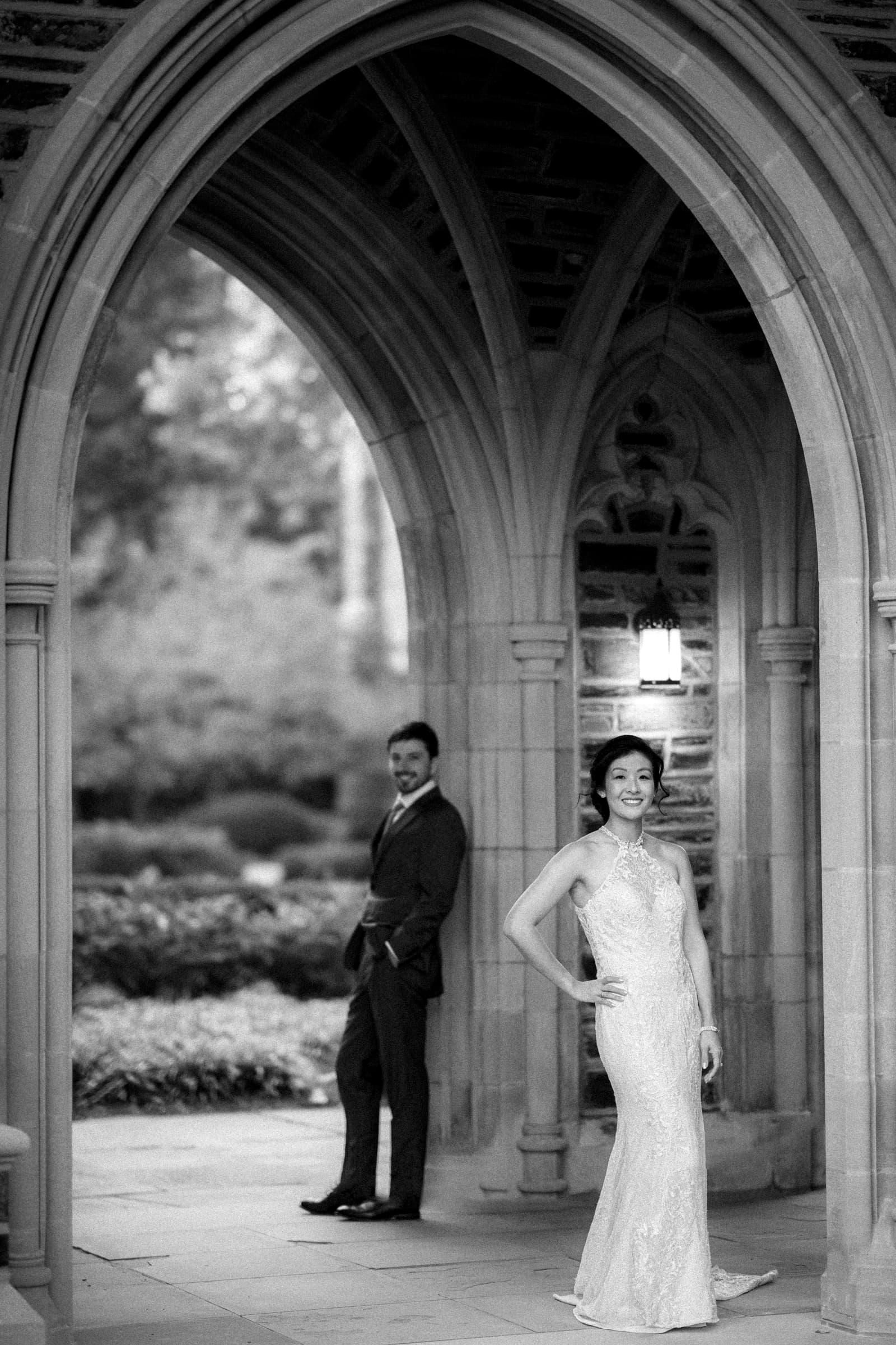 A bride in a long, elegant gown poses under a stone archway, smiling. In the background, her groom in a suit stands casually, leaning against the arch. The market hall wedding scene is outdoors, surrounded by greenery and soft natural light.