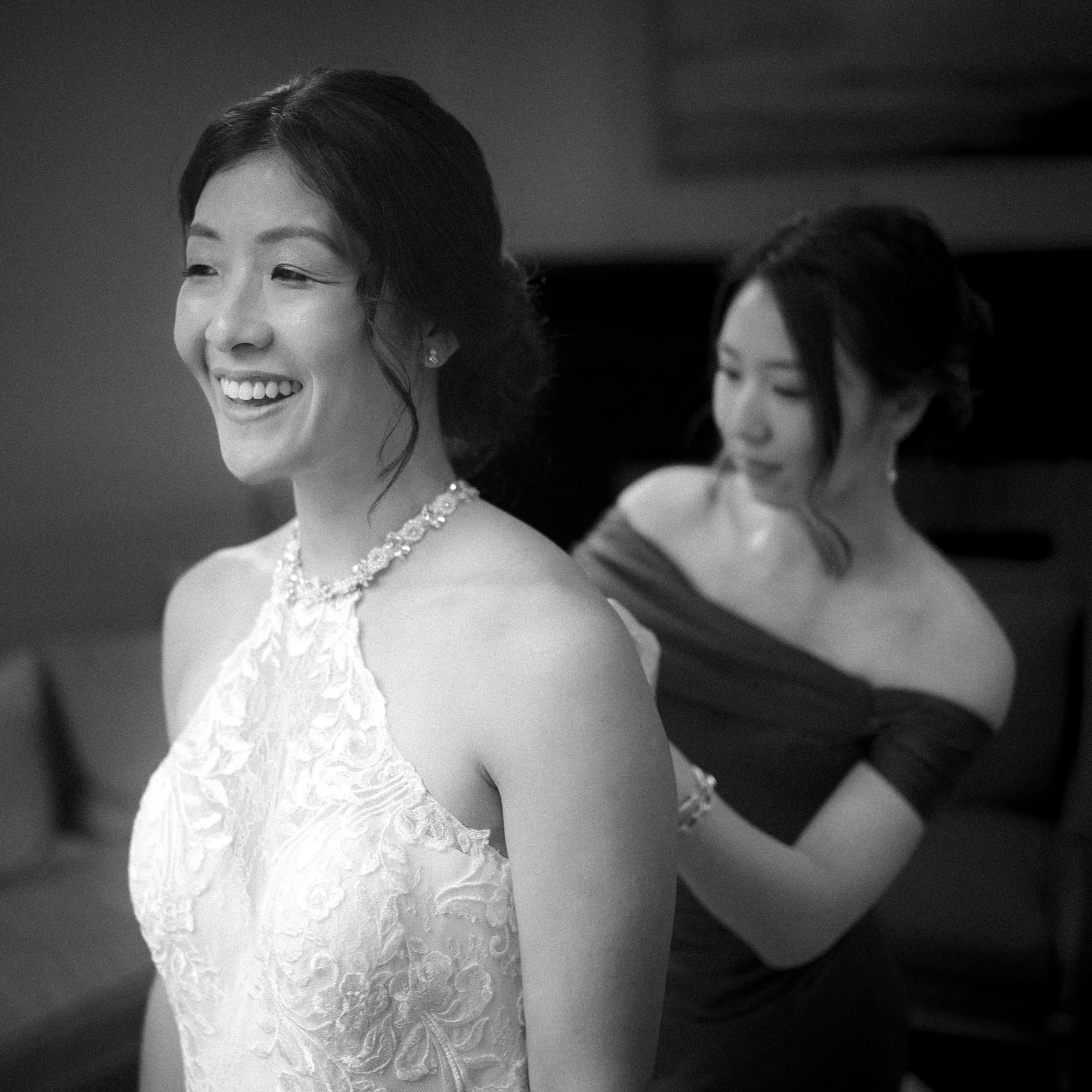 A bride beams with joy in her wedding dress as another woman helps adjust the back, both filled with happiness in the softly lit setting of a market hall wedding.