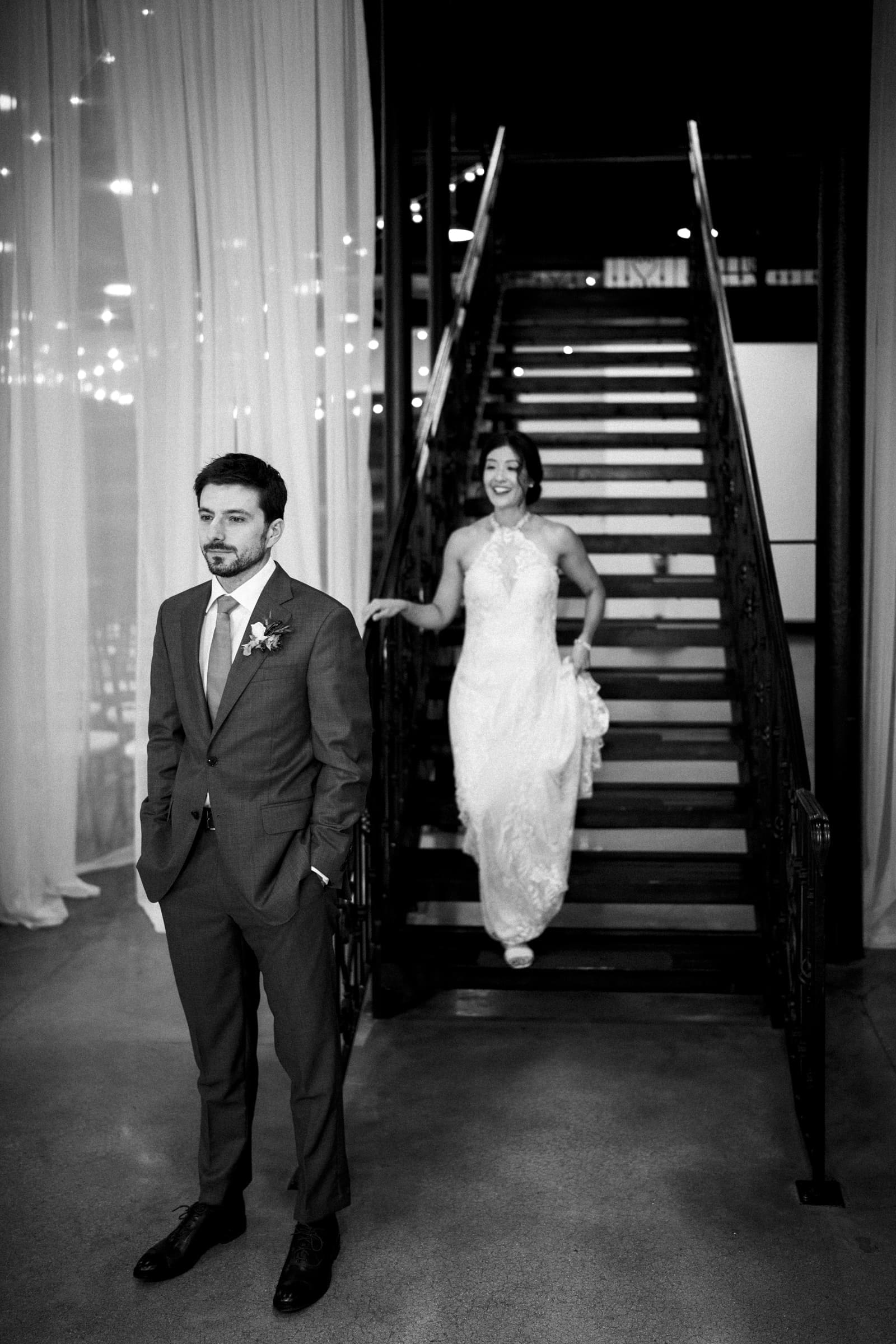 A black and white photo captures a groom in a suit, hands in pockets, looking forward. Behind him, a bride in a lace gown descends the staircase of the market hall venue, smiling. The softly lit scene is adorned with glowing string lights in the background.