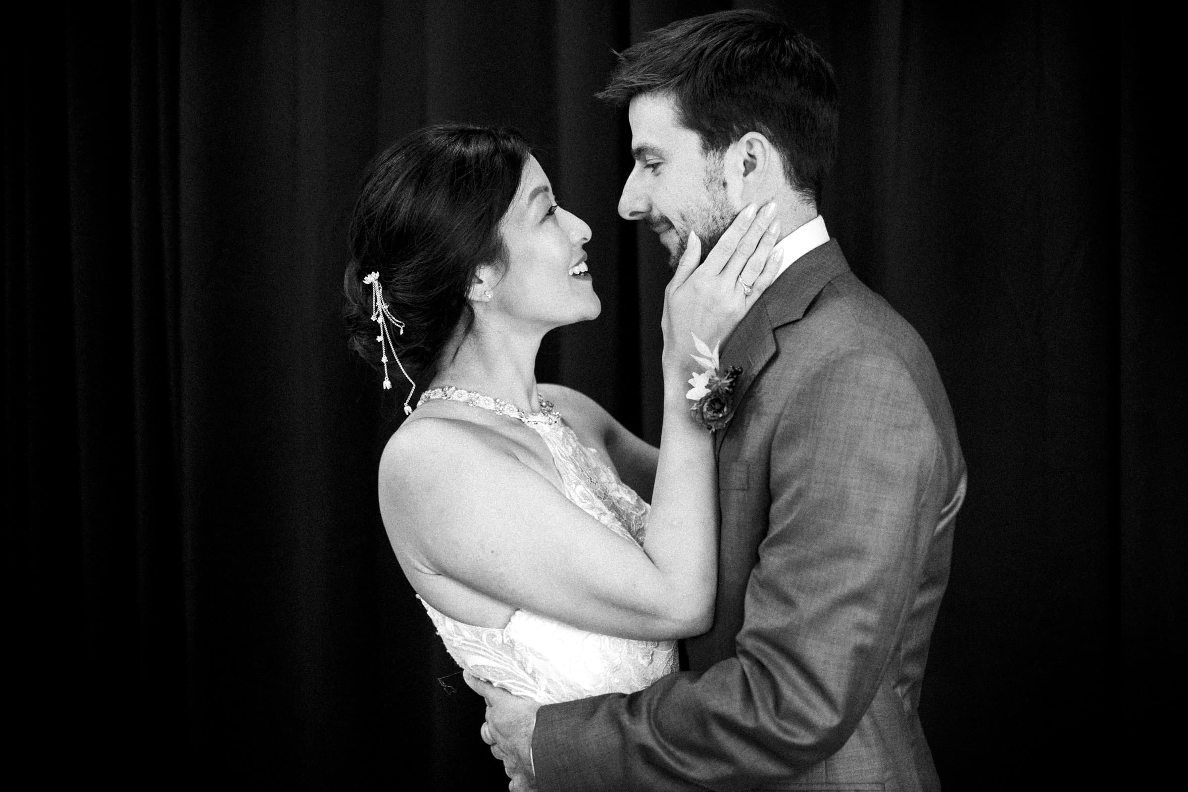 A black and white image of a smiling couple on their market hall wedding day. The bride, in a lace dress, holds the groom’s face gently with her hands. The groom, in a suit, embraces her waist. Both look into each other's eyes against a dark backdrop.