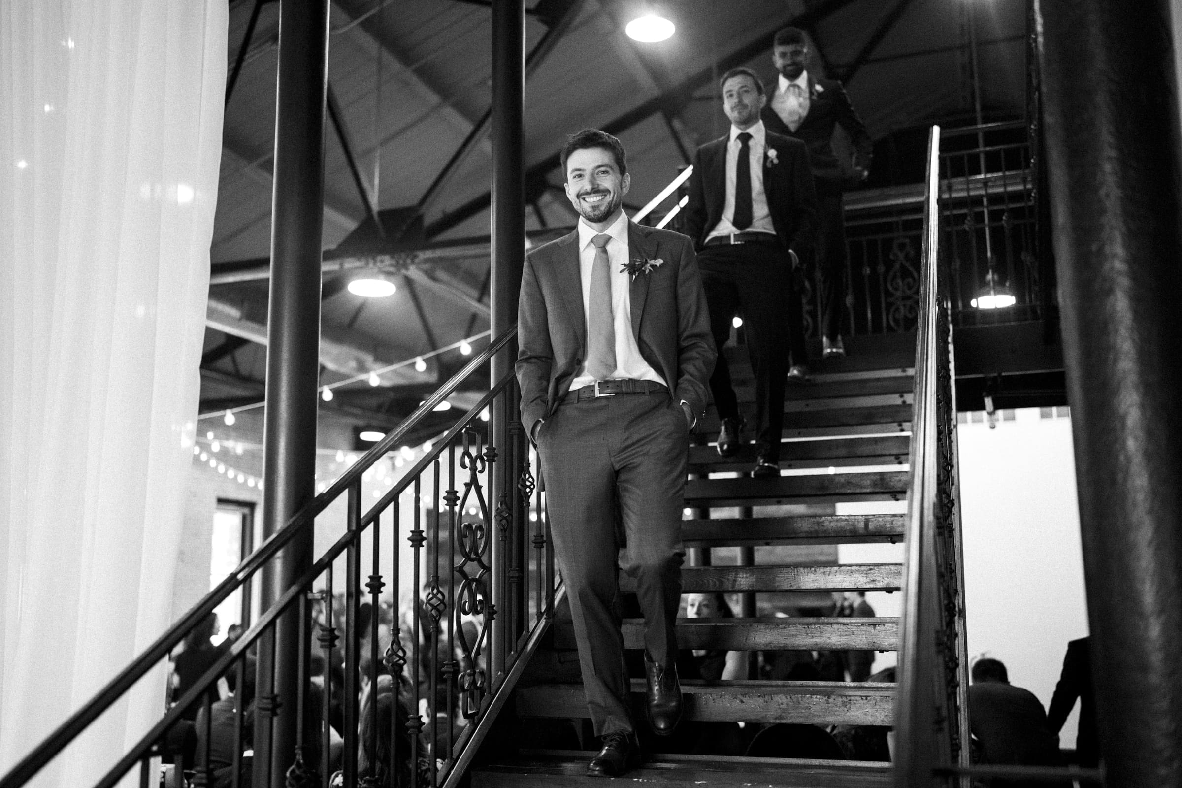 Three men in suits walk down a staircase at a market hall wedding, smiling and engaged in conversation. The setting features decorative lighting and a high ceiling, creating a festive atmosphere. The black-and-white image captures the timeless elegance of the celebration.