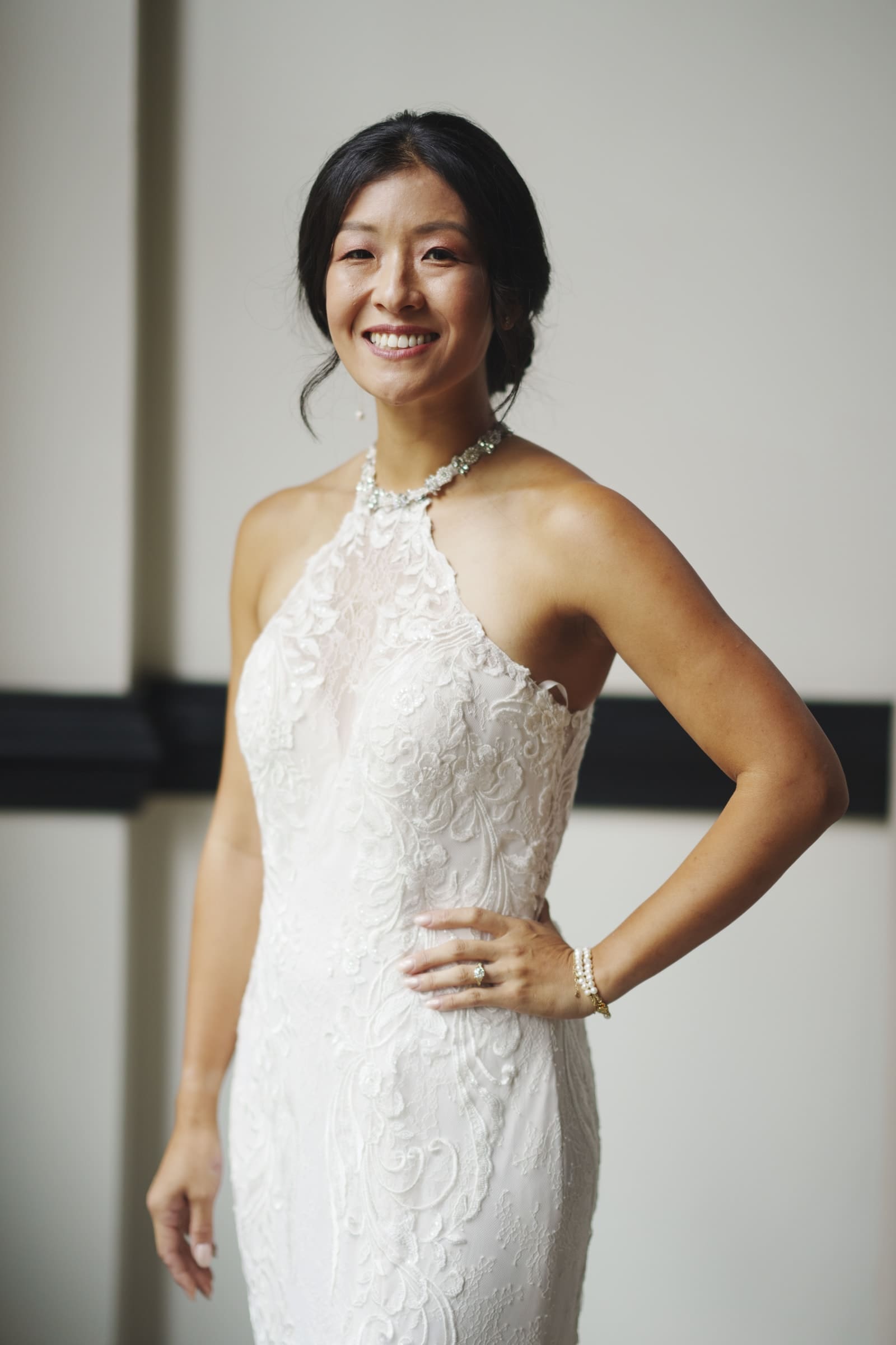 A woman in a sleeveless, lace bridal gown poses with a hand on her hip, smiling against the softly lit backdrop of a market hall wedding. Her hair is styled up, and she wears a necklace and bracelet, perfectly complementing the elegant atmosphere.