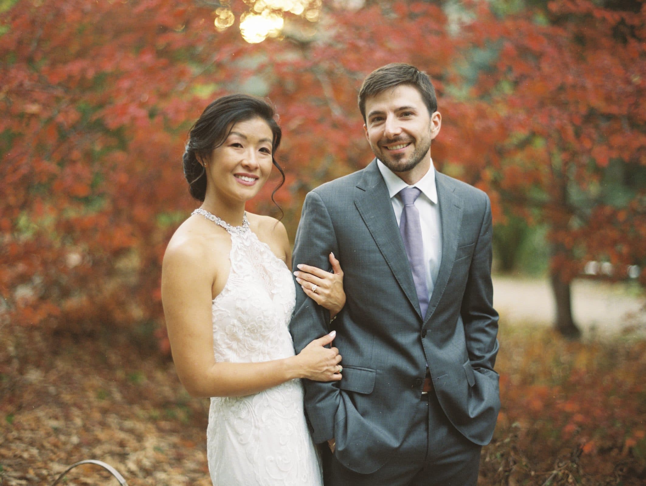 A smiling couple poses outdoors in front of vibrant autumn foliage, reminiscent of a picturesque market hall wedding. The woman is in a white lace dress, and the man sports a gray suit with a lavender tie. The serene setting bathes them in warm, natural light.