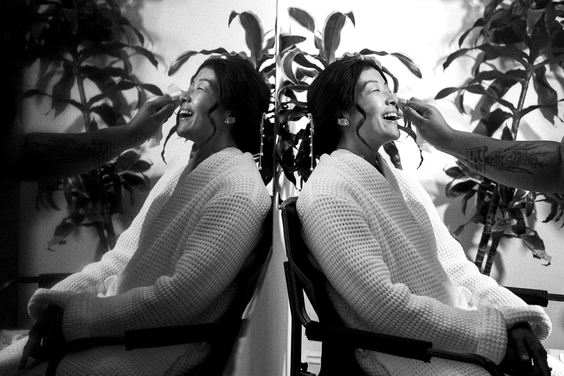 A bride in a robe sits smiling as her makeup is meticulously applied, preparing for her market hall wedding. She is seated in front of a large mirror, reflecting her radiant image. A plant graces the background, adding a touch of nature to the vibrant scene.