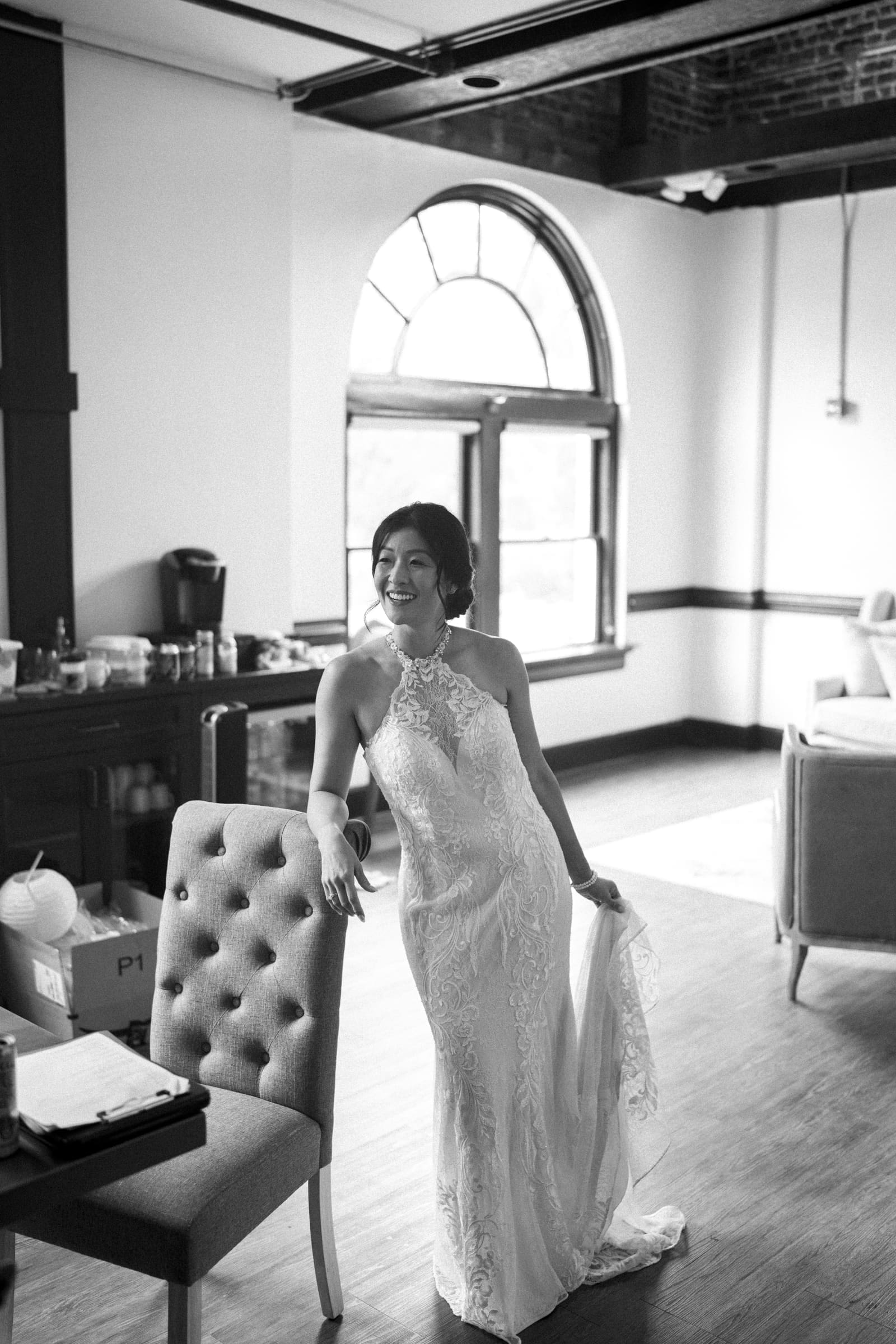 A bride in a lace wedding dress poses in a sunlit room, evoking the charm of a market hall wedding. She stands next to a tufted chair, holding her gown. The room features arched windows and exposed brick, creating a warm, elegant atmosphere.
