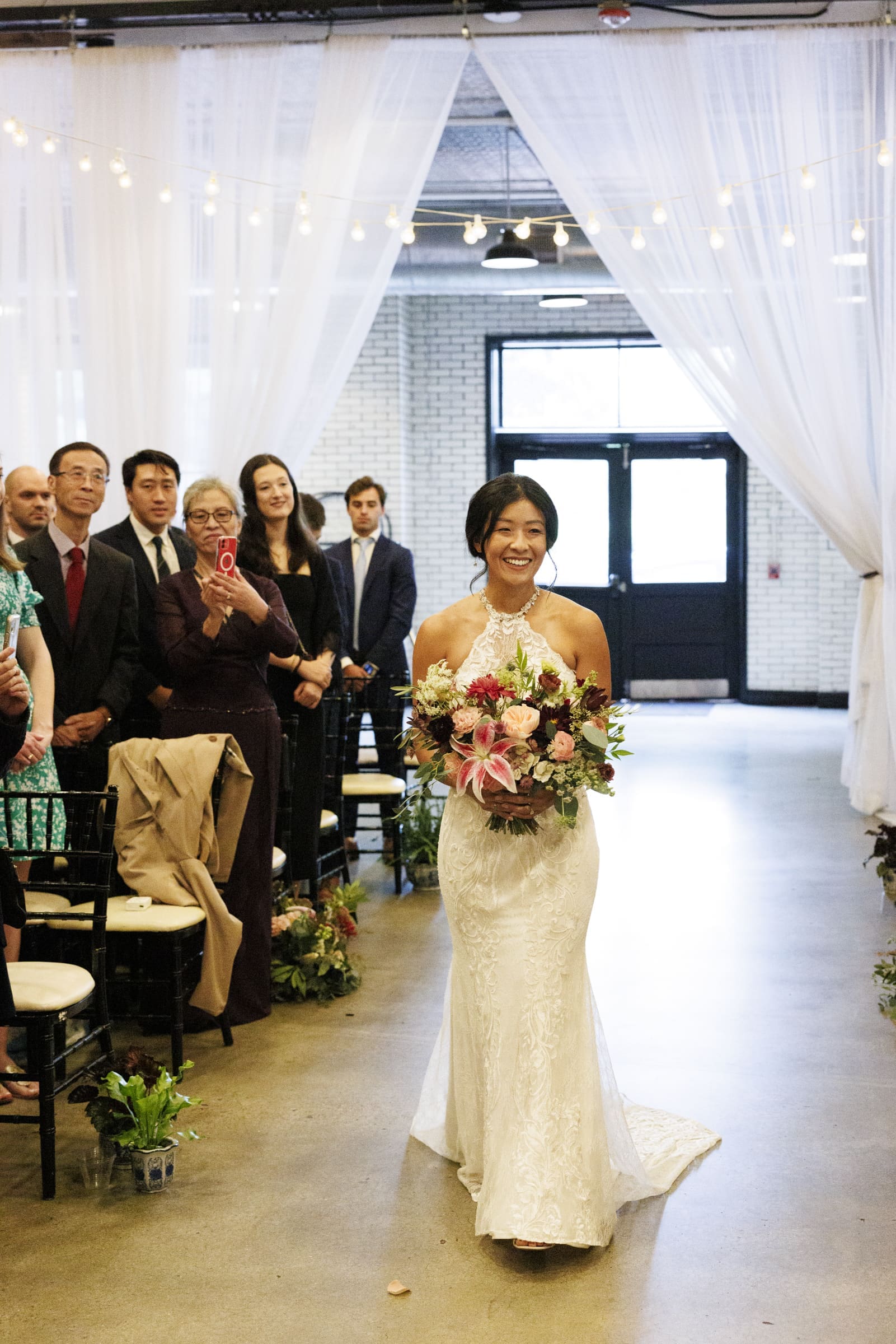 A bride in a white gown walks down the aisle of the market hall, holding a colorful bouquet. Guests stand on either side, capturing the moment. The venue is adorned with sheer white curtains and string lights, adding charm to this unique wedding setting.