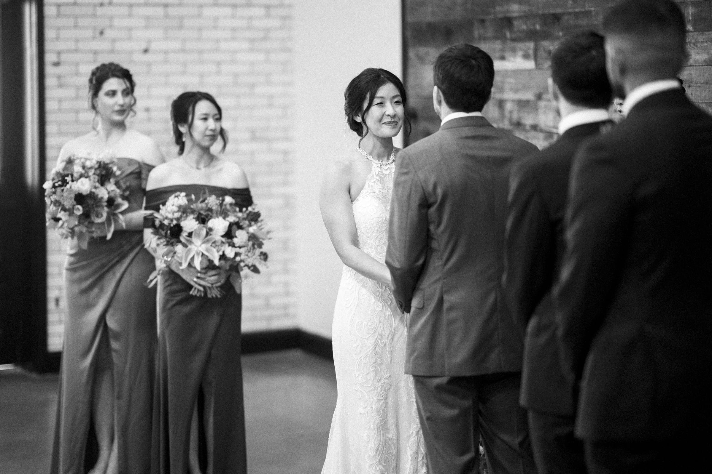 A bride and groom exchange vows in a market hall wedding ceremony. The bride smiles while holding the groom's hands. Bridesmaids stand in the background, holding bouquets. The setting is indoors with a brick wall, and the image is elegantly captured in black and white.