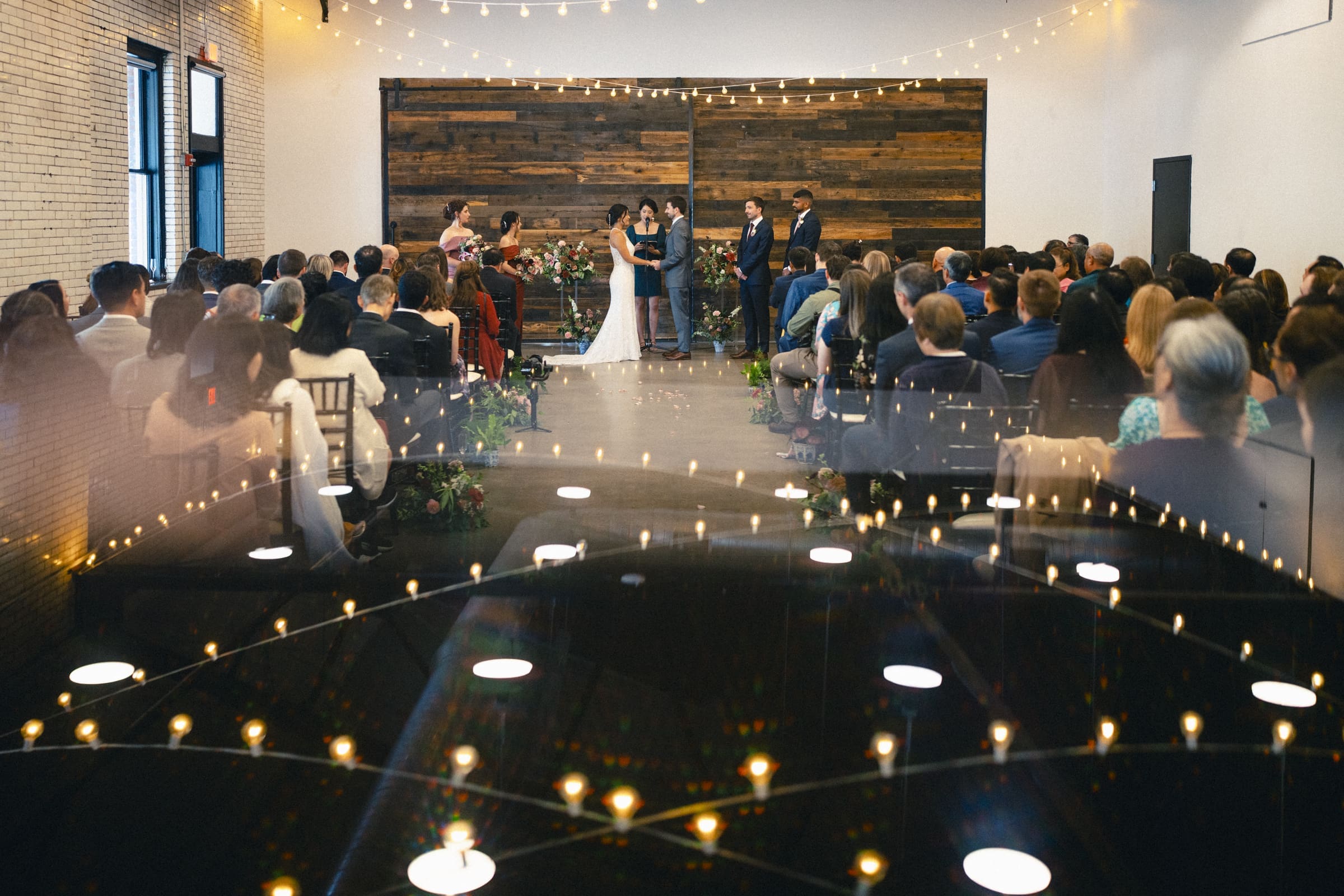 A market hall wedding unfolds in a rustic venue with wooden walls and string lights. The couple stands at the altar with the officiant and bridesmaids, surrounded by seated guests. Bright lights create a soft, reflective ambiance across the room.