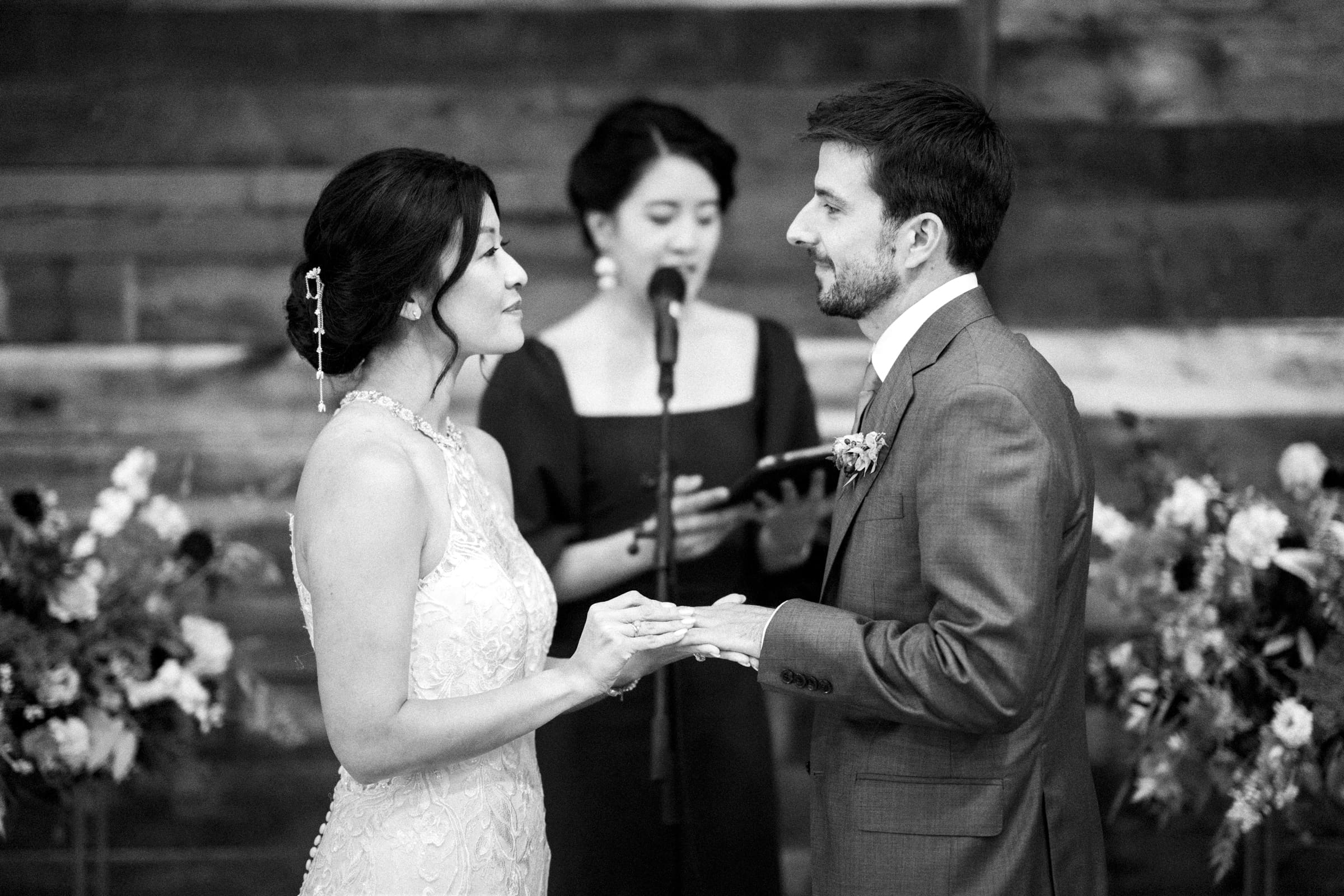 A bride and groom hold hands during their market hall wedding ceremony. They face each other as a person stands behind them, speaking into a microphone. The background features wooden walls and floral arrangements, enriching the setting. The image is captured in timeless black and white.