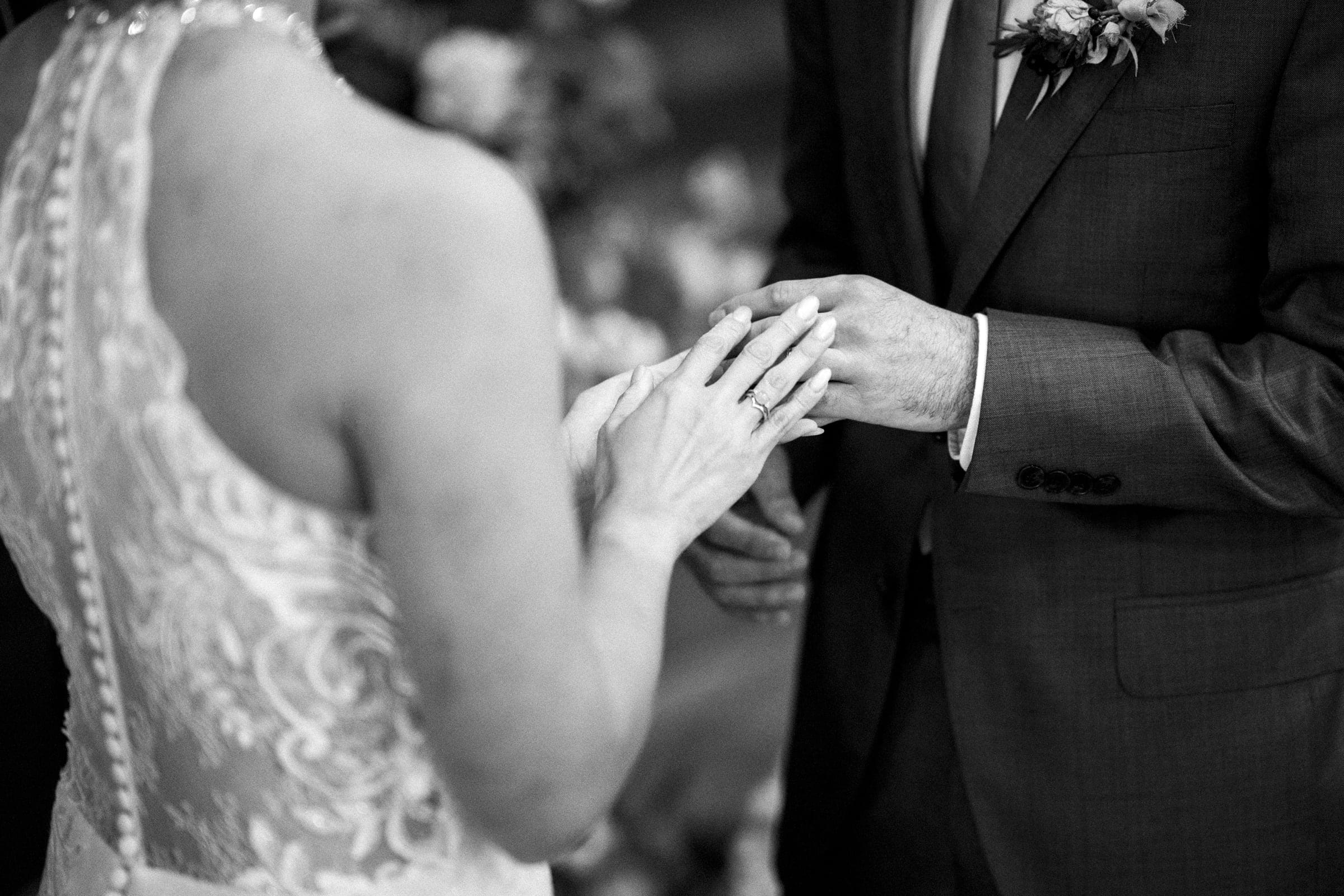 A black-and-white close-up captures the intimate exchange at a market hall wedding. The bride, in a lace dress, delicately places a ring on the groom's finger. He wears a suit with a boutonniere, emphasizing the tender focus on their hands during this cherished moment.