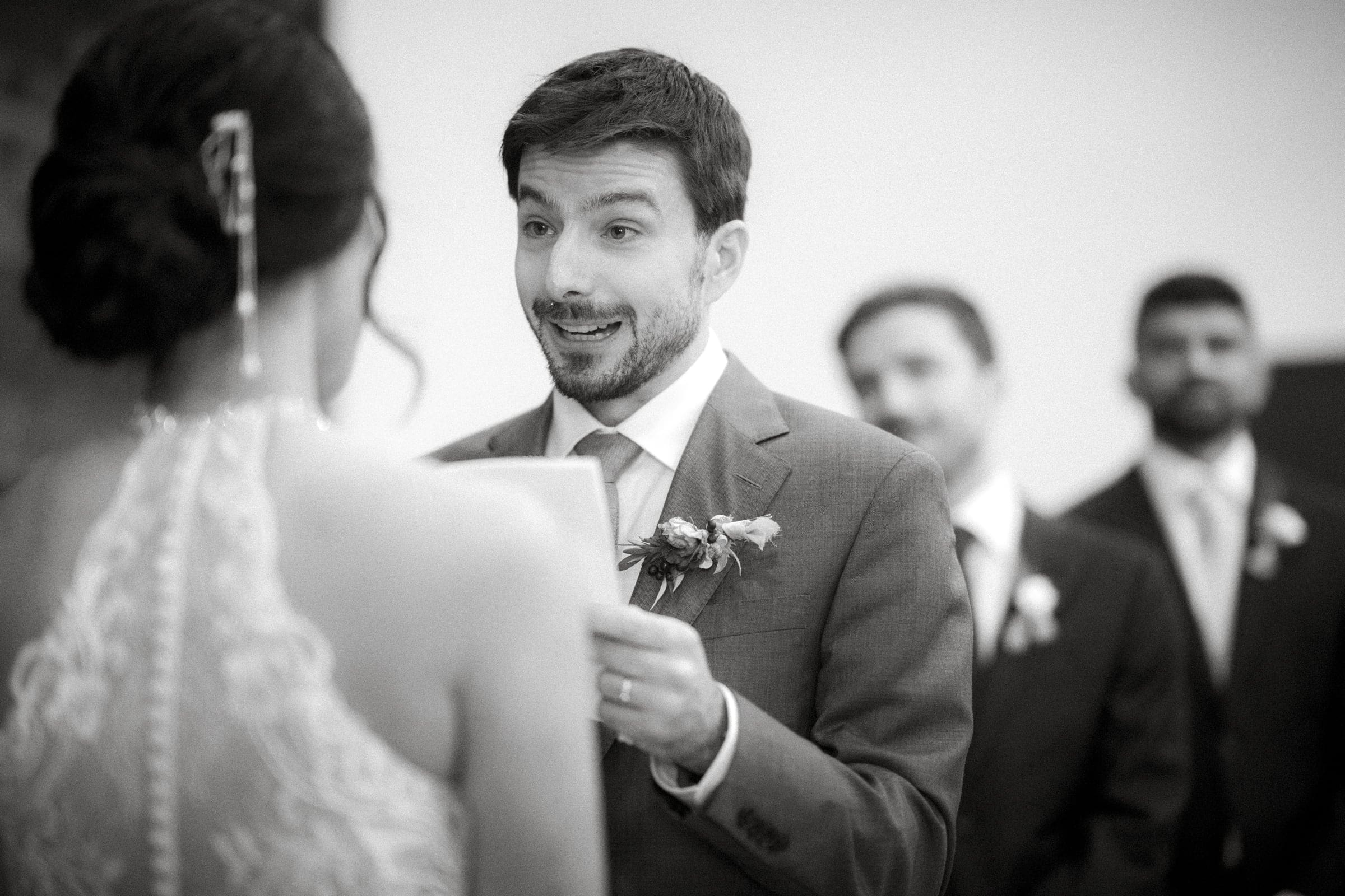A man in a suit, holding a piece of paper, speaks animatedly to a woman in a detailed dress at the market hall wedding. Three men in suits stand in the background. The black-and-white scene captures an emotional moment during the ceremony.