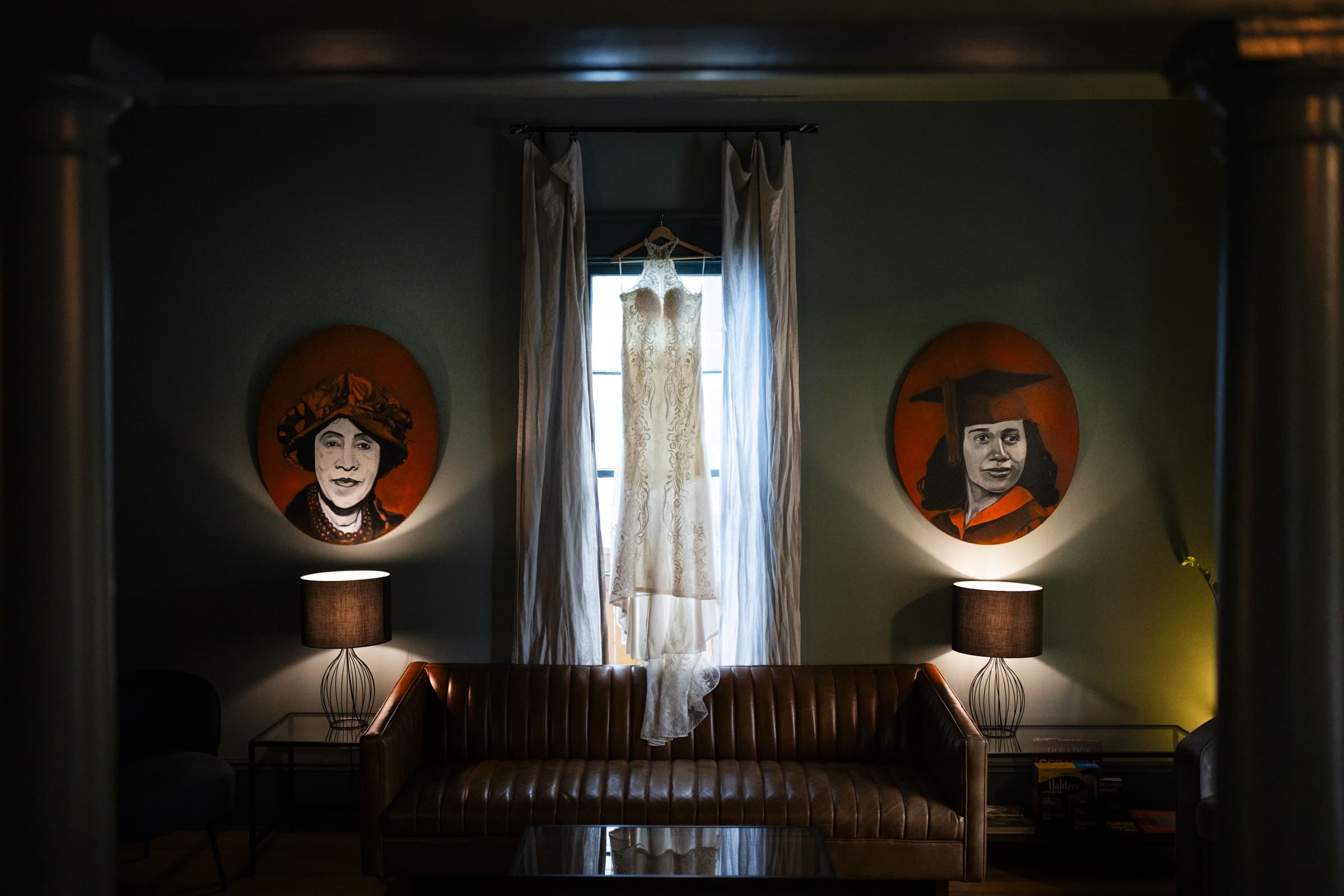 A white lace wedding dress is elegantly displayed in front of a window with light curtains at the market hall wedding venue. It hangs between two round portraits, one of a woman wearing a hat and another of a woman in a graduation cap. Two lit table lamps flank a brown leather sofa.
