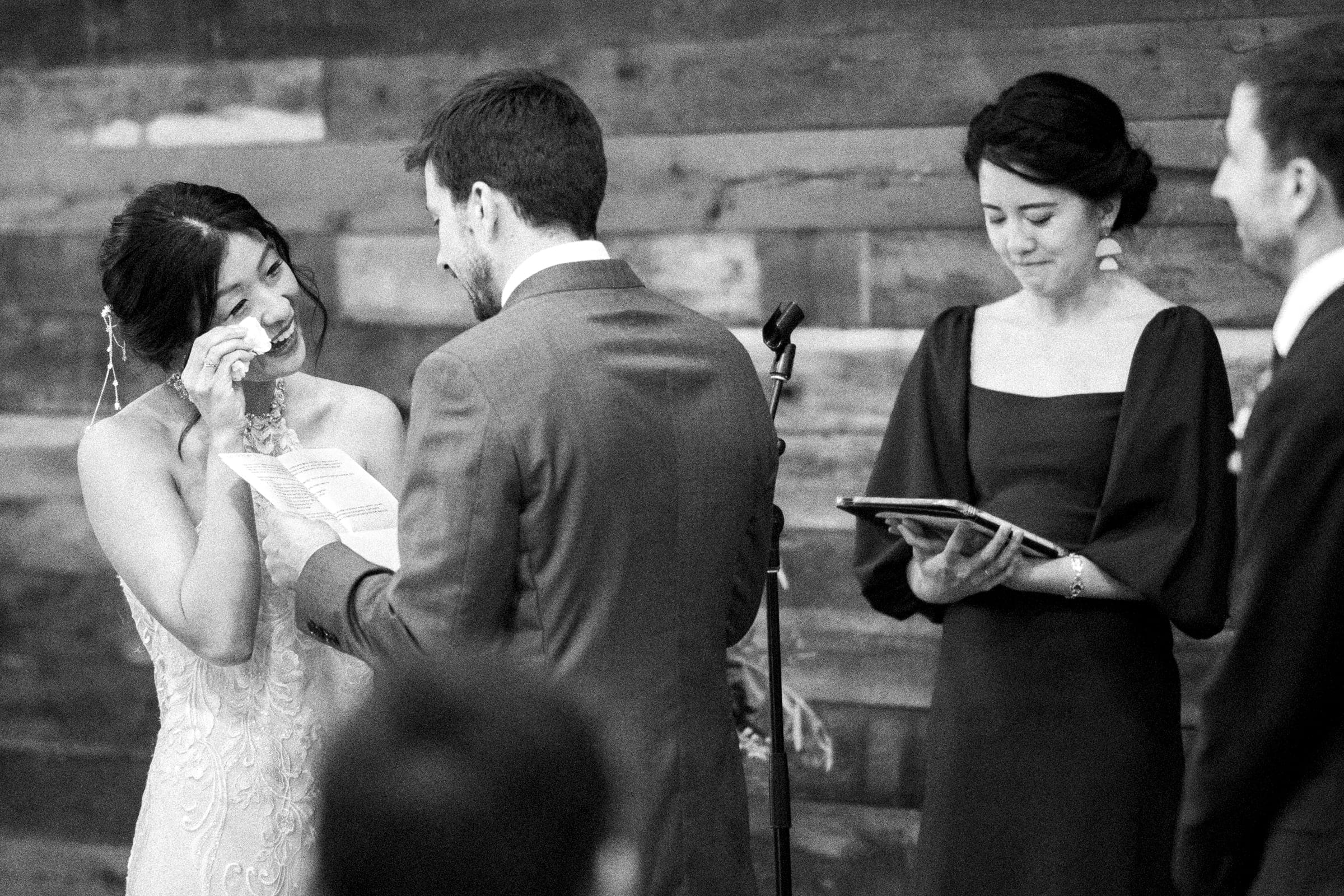 A bride wipes her tears while reading vows during an intimate market hall wedding ceremony. The groom smiles, holding his vows, as a person officiates with a smile, holding a book. Another guest watches amidst the rustic wooden backdrop.