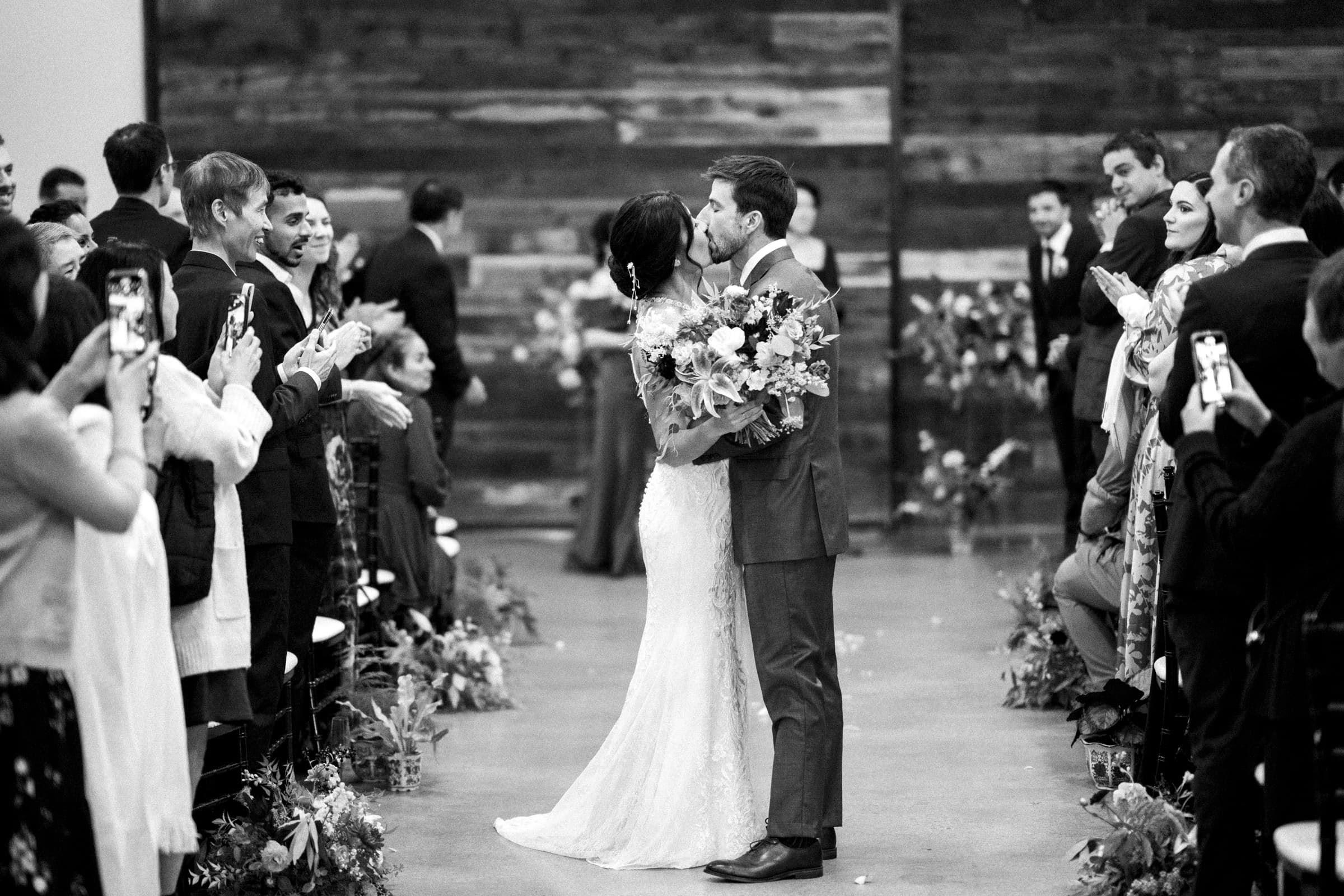 At the end of the aisle in a market hall wedding, a bride and groom share a kiss. Guests on either side clap and capture photos against the rustic wooden backdrop adorned with flowers. The timeless black-and-white image beautifully encapsulates this special moment.