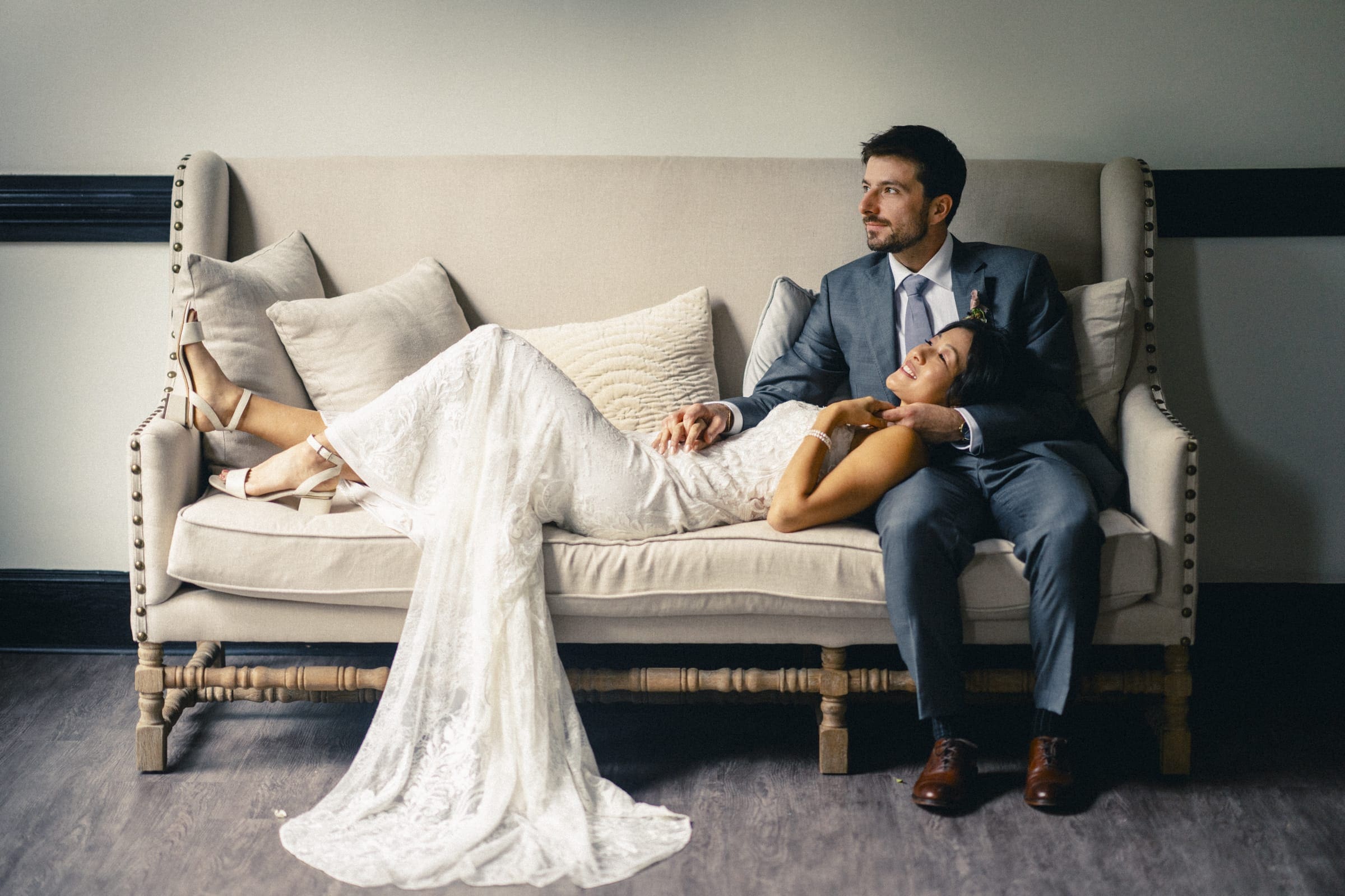 A couple is sitting on a beige couch in a market hall. The woman, in a white wedding dress, reclines with her head on the man's lap. The man, in a gray suit, gazes into the distance, while she appears relaxed amid the cushions.