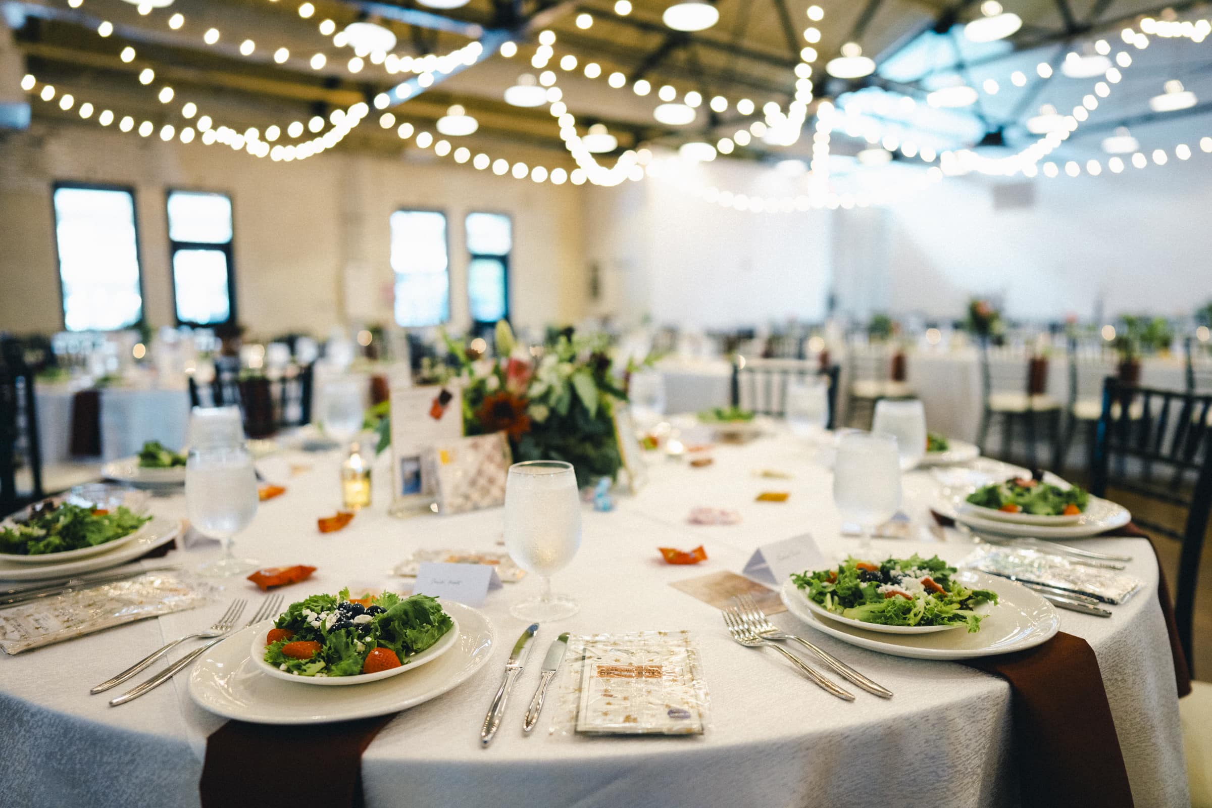 The beautifully decorated market hall features round tables set for a wedding. Each table boasts white tablecloths, salads on plates, and elegant place settings, while string lights craft a warm, inviting atmosphere.