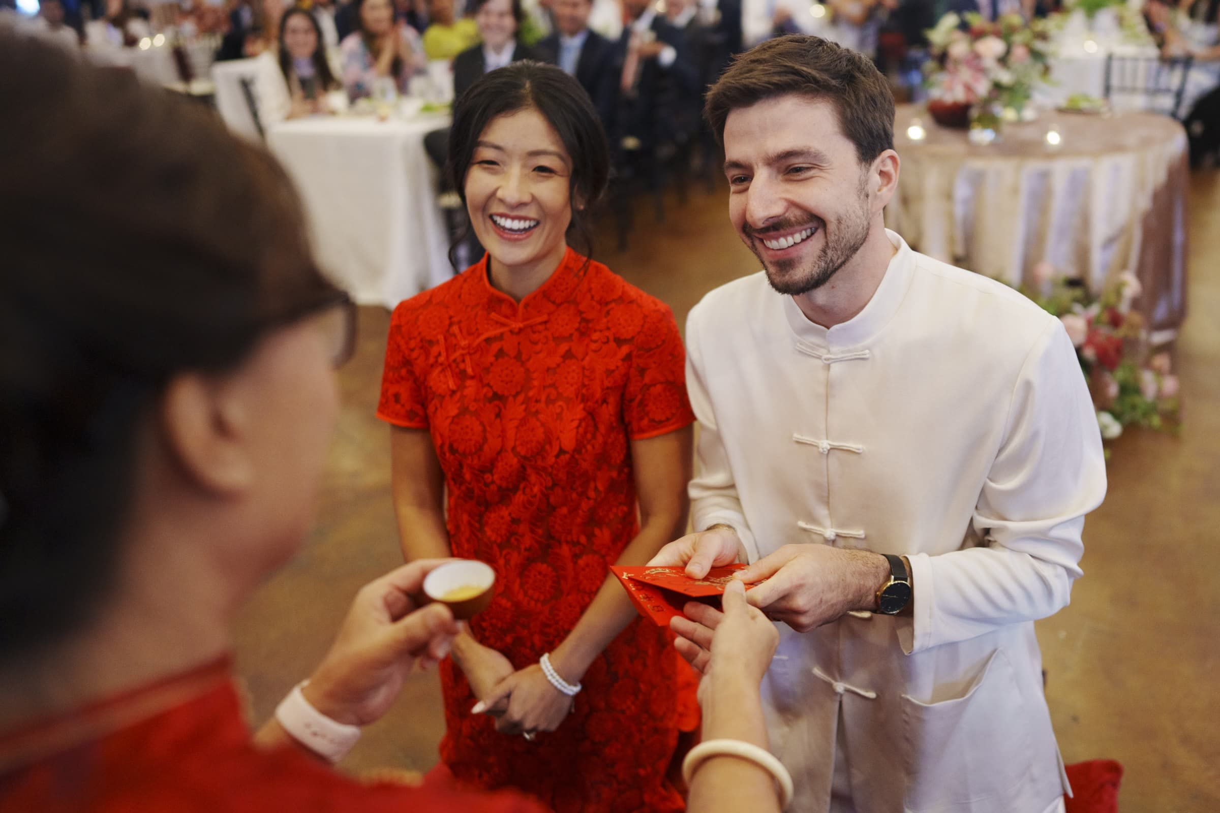 A couple in traditional attire smiles during a tea ceremony at a festive market hall wedding. The woman wears a red dress, and the man dons a white outfit. They hand a red envelope to someone in front of them as guests enjoy the celebration at tables in the background.
