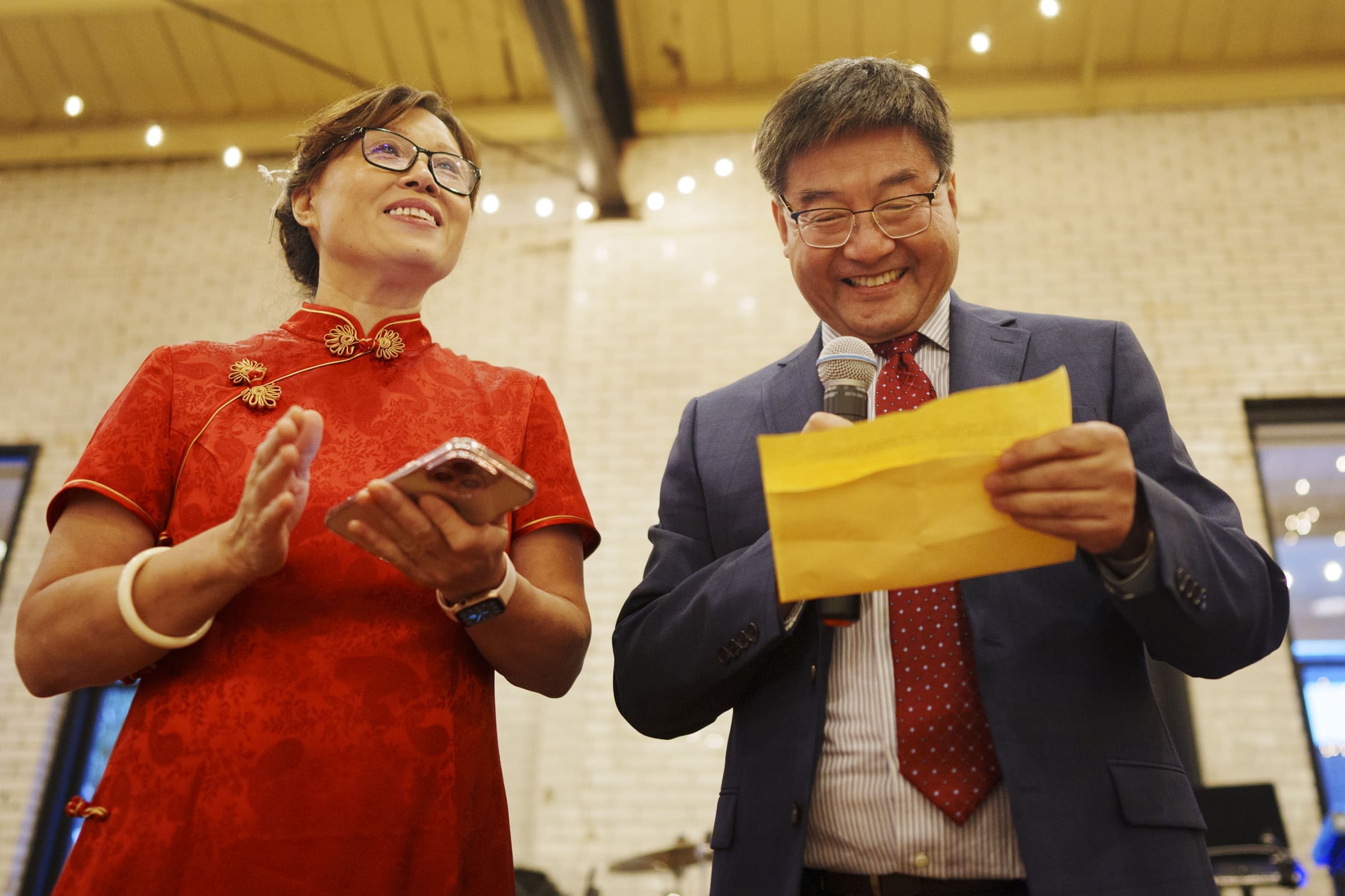 A woman in a red dress holds a smartphone, smiling and clapping as a man in a suit reads from yellow paper. The scene unfolds in a well-lit room adorned with string lights. They are celebrating at what seems to be an enchanting market hall wedding, both clearly enjoying the moment.