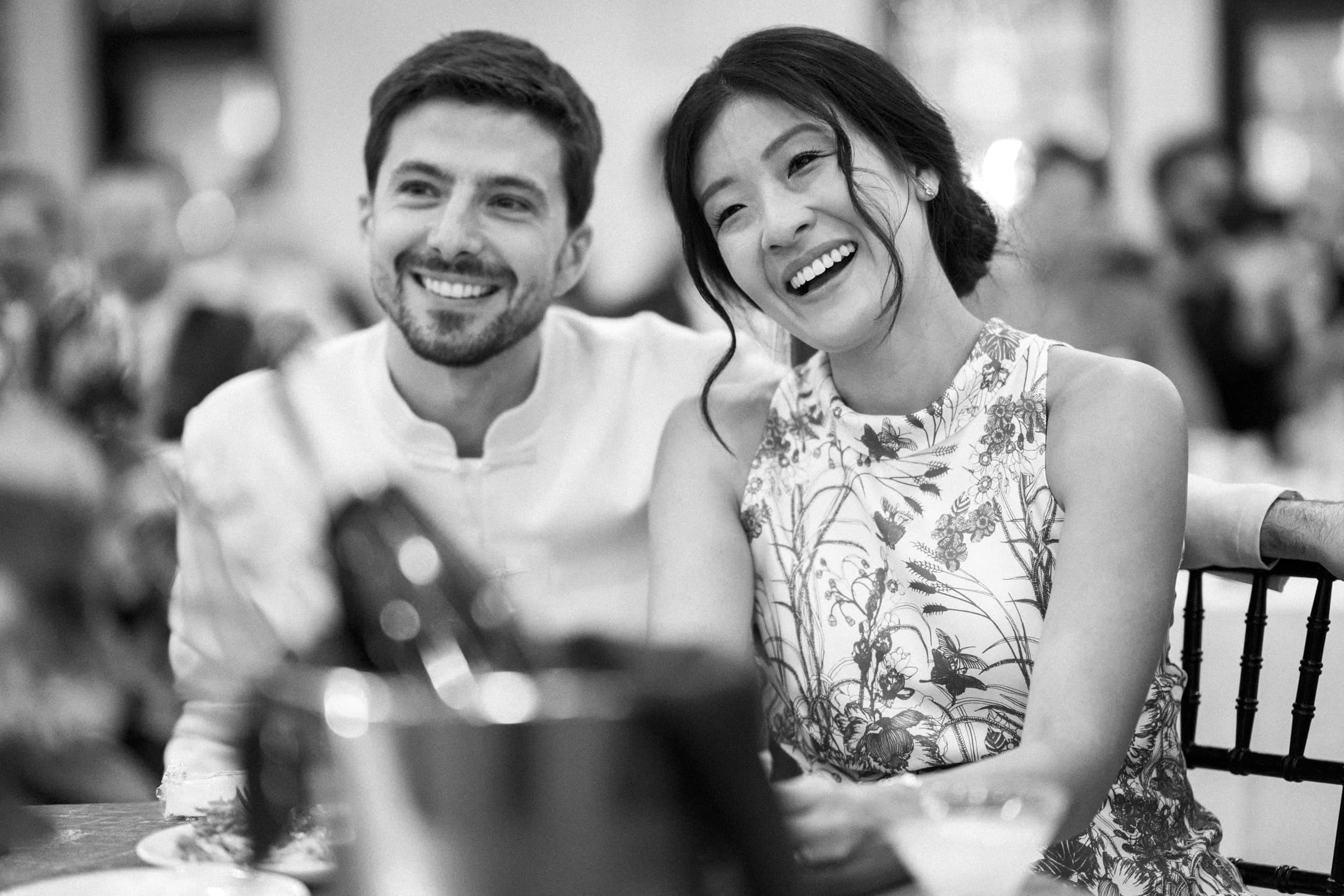 A black and white photo captures a smiling couple at a market hall wedding. The woman wears a floral dress, and the man is in a light-colored shirt. They sit closely, looking content and relaxed, with blurred people and decorations in the background.