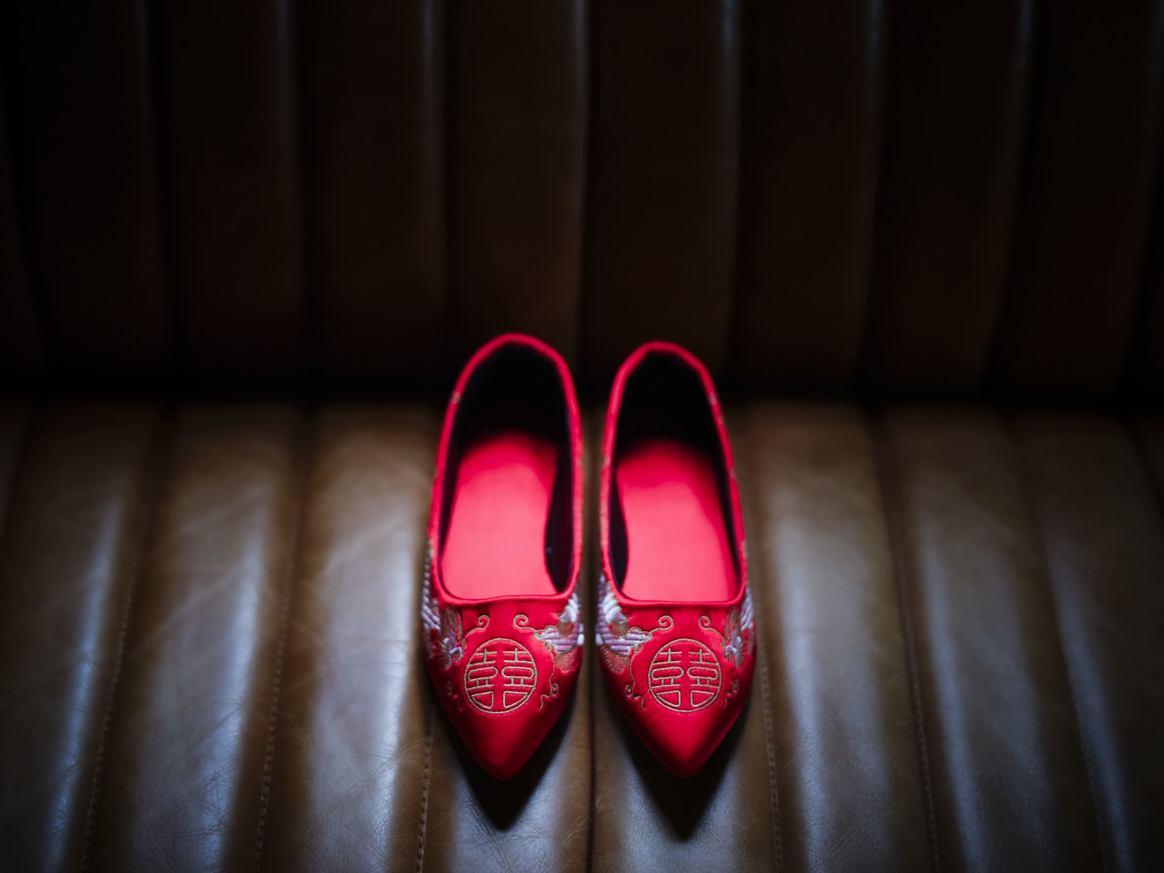 A pair of red, embroidered shoes with traditional Chinese symbols rests against a dark, textured background, reminiscent of a market hall's vibrant atmosphere. The intricate design and color echo the celebratory spirit one might find at a wedding.